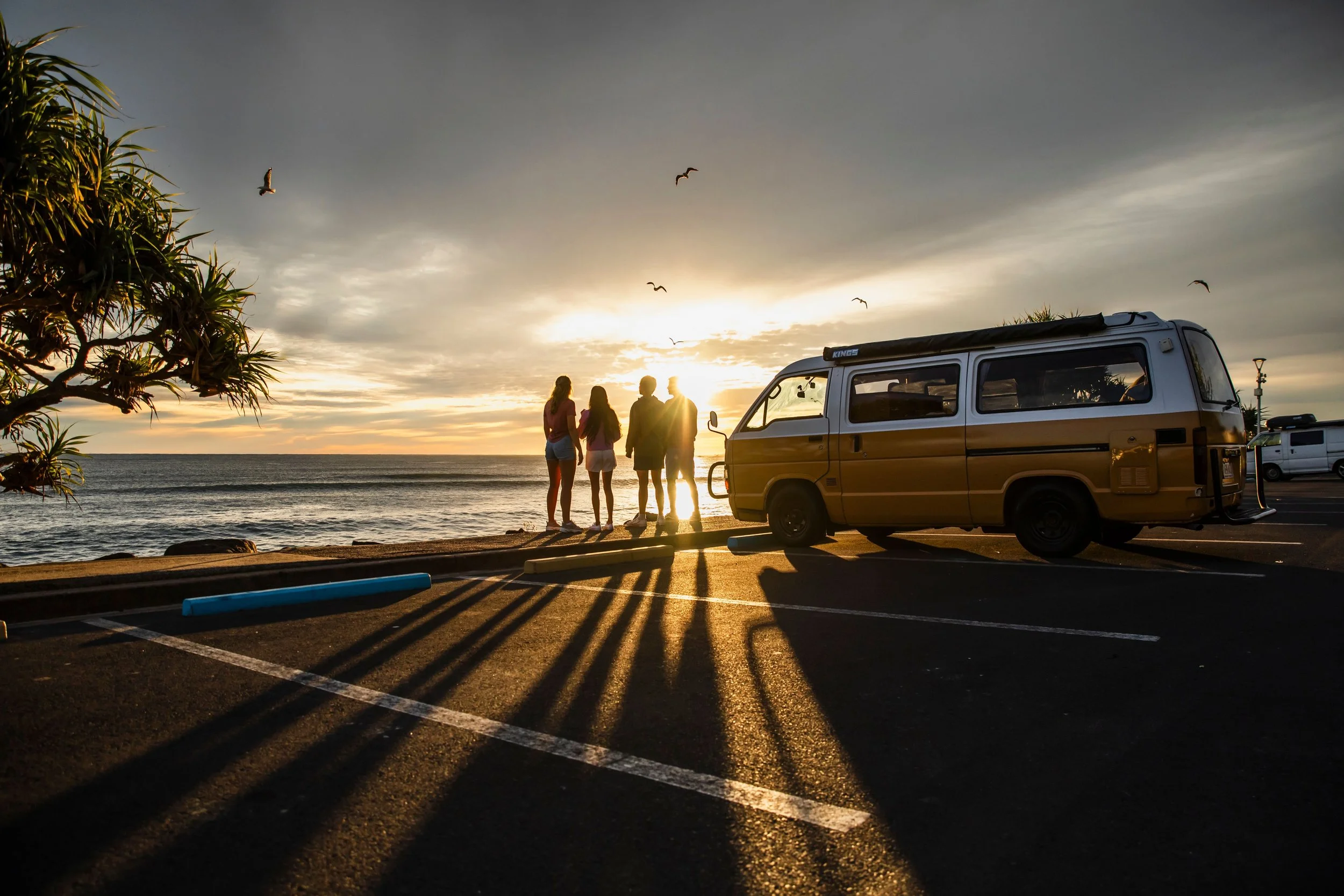 A small yellow camper van driving on a roundabout in a suburban area with palm trees and shops in the background under a partly cloudy sky.