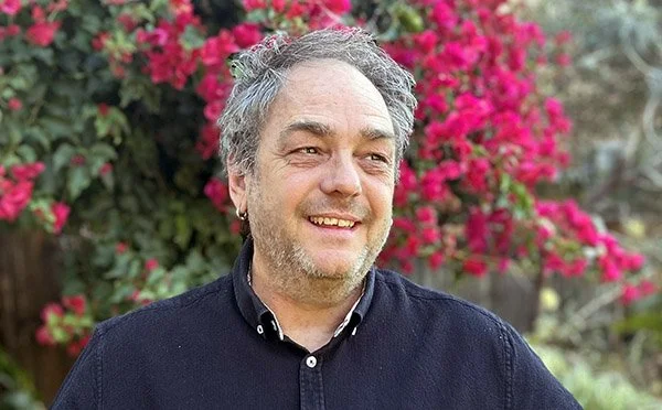 A man with gray hair and a beard smiling outdoors, with pink flowers and greenery in the background.