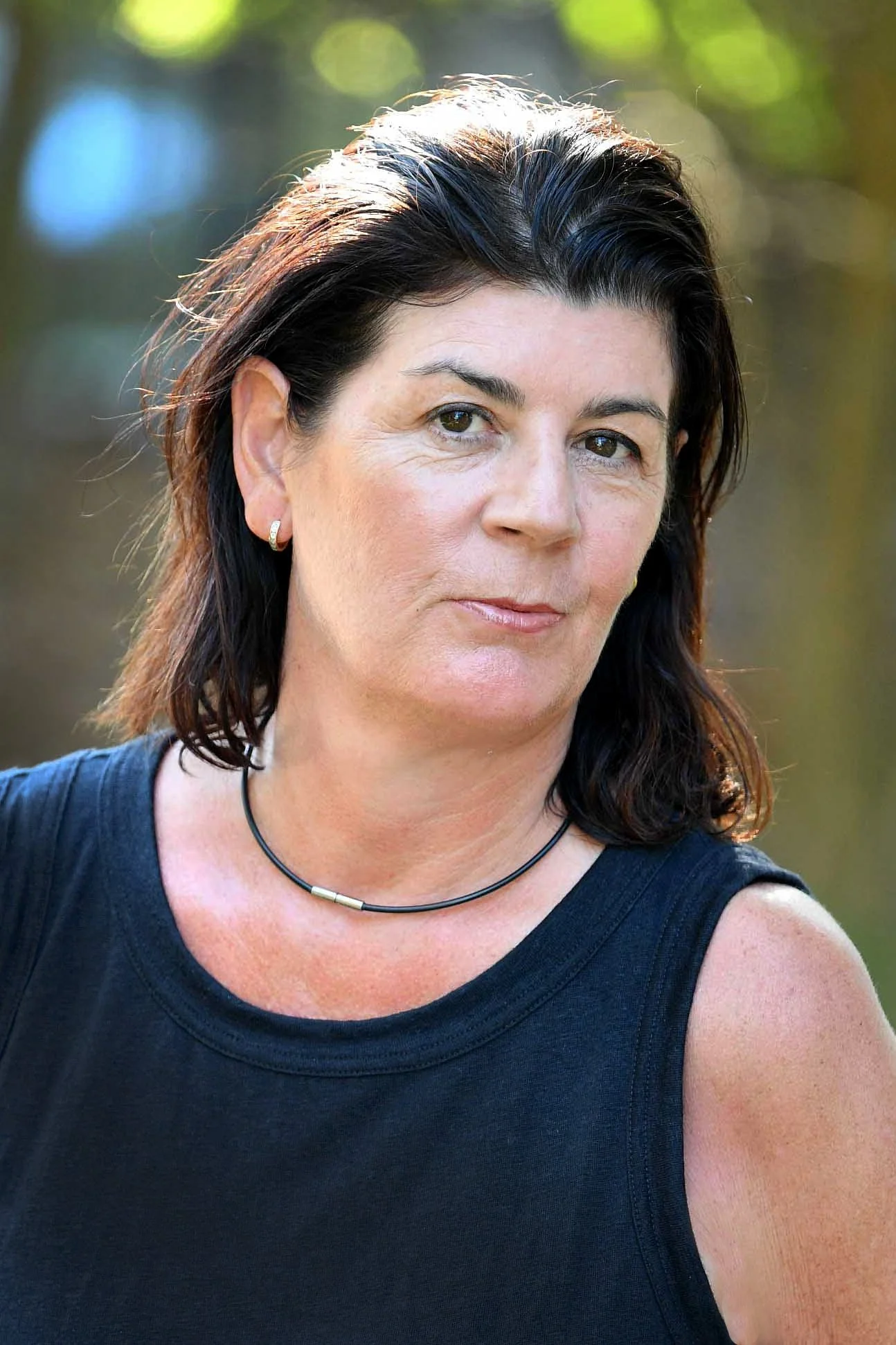 A middle-aged woman with dark brown hair and light skin, wearing a black sleeveless top, a black choker necklace, and small hoop earrings, outdoors with blurred trees in the background.