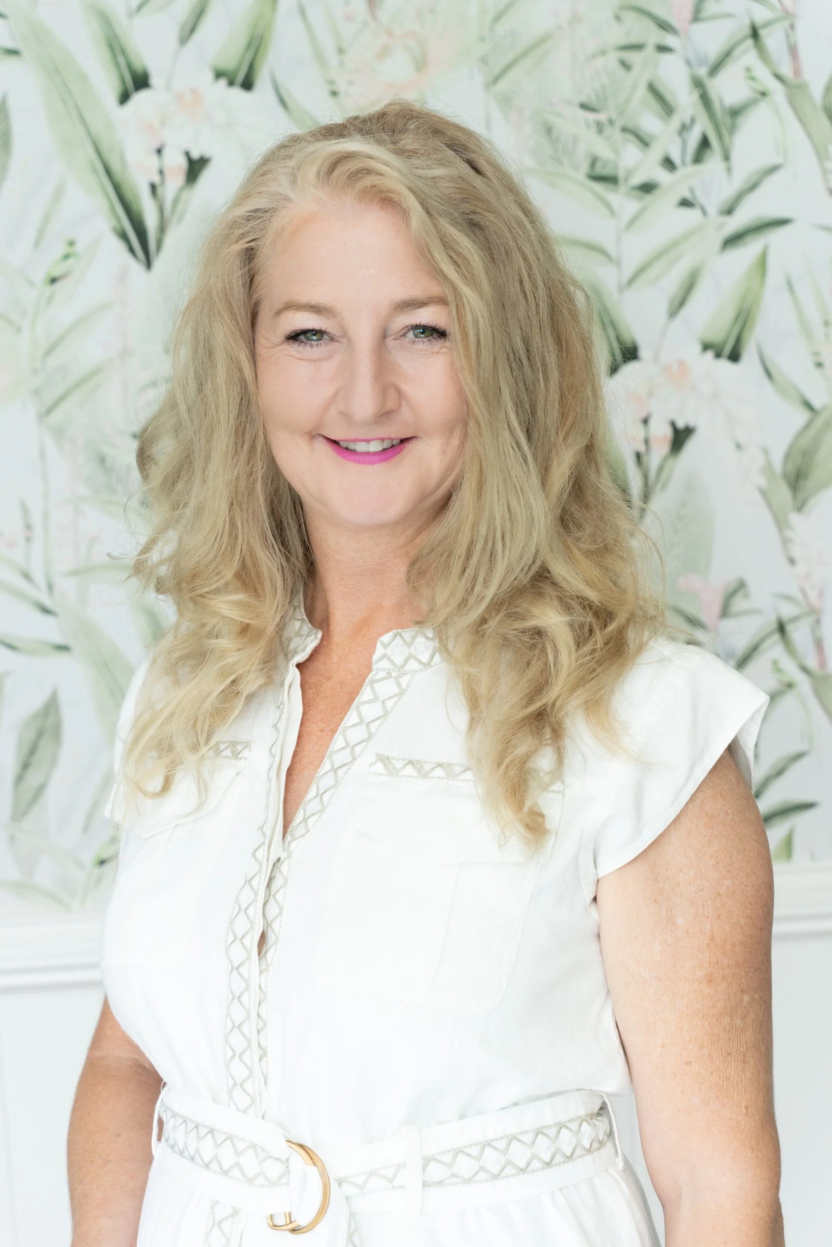 Portrait of a smiling woman with blonde curly hair, wearing a white dress with a belt, standing in front of a floral wallpaper.