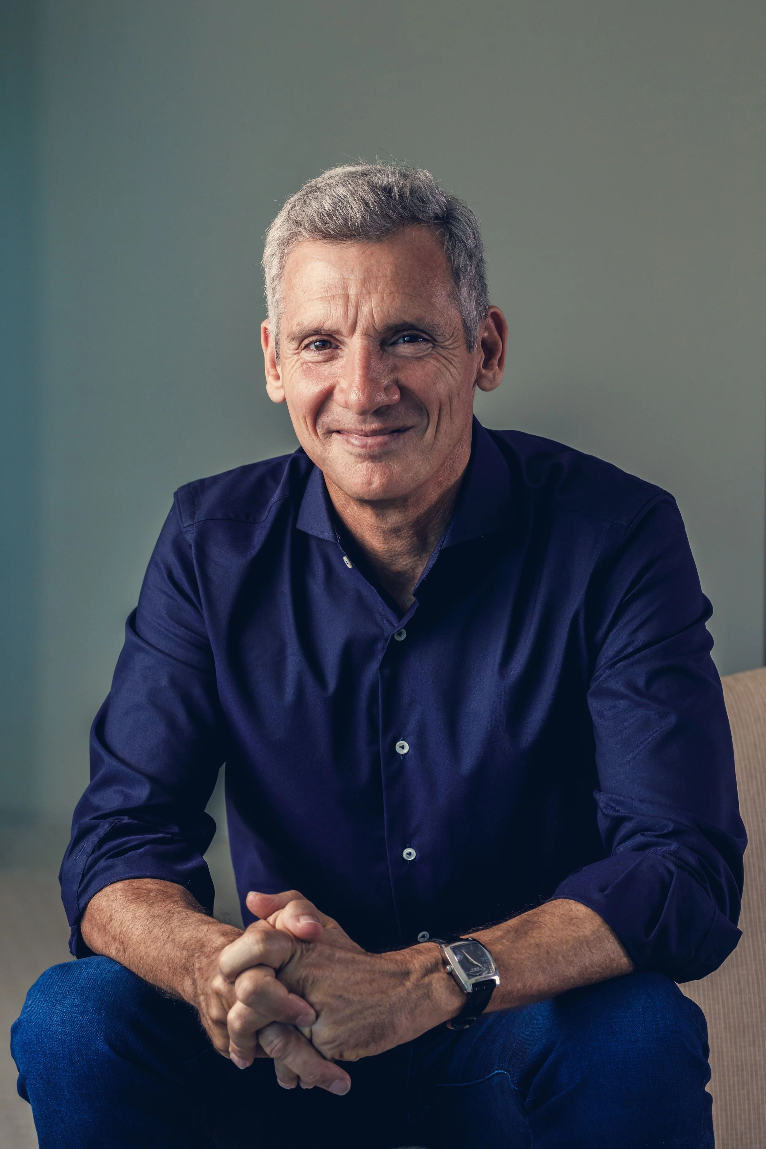 A middle-aged man with gray hair, wearing a dark blue button-up shirt and a watch, sitting with hands clasped, smiling in a professional setting.