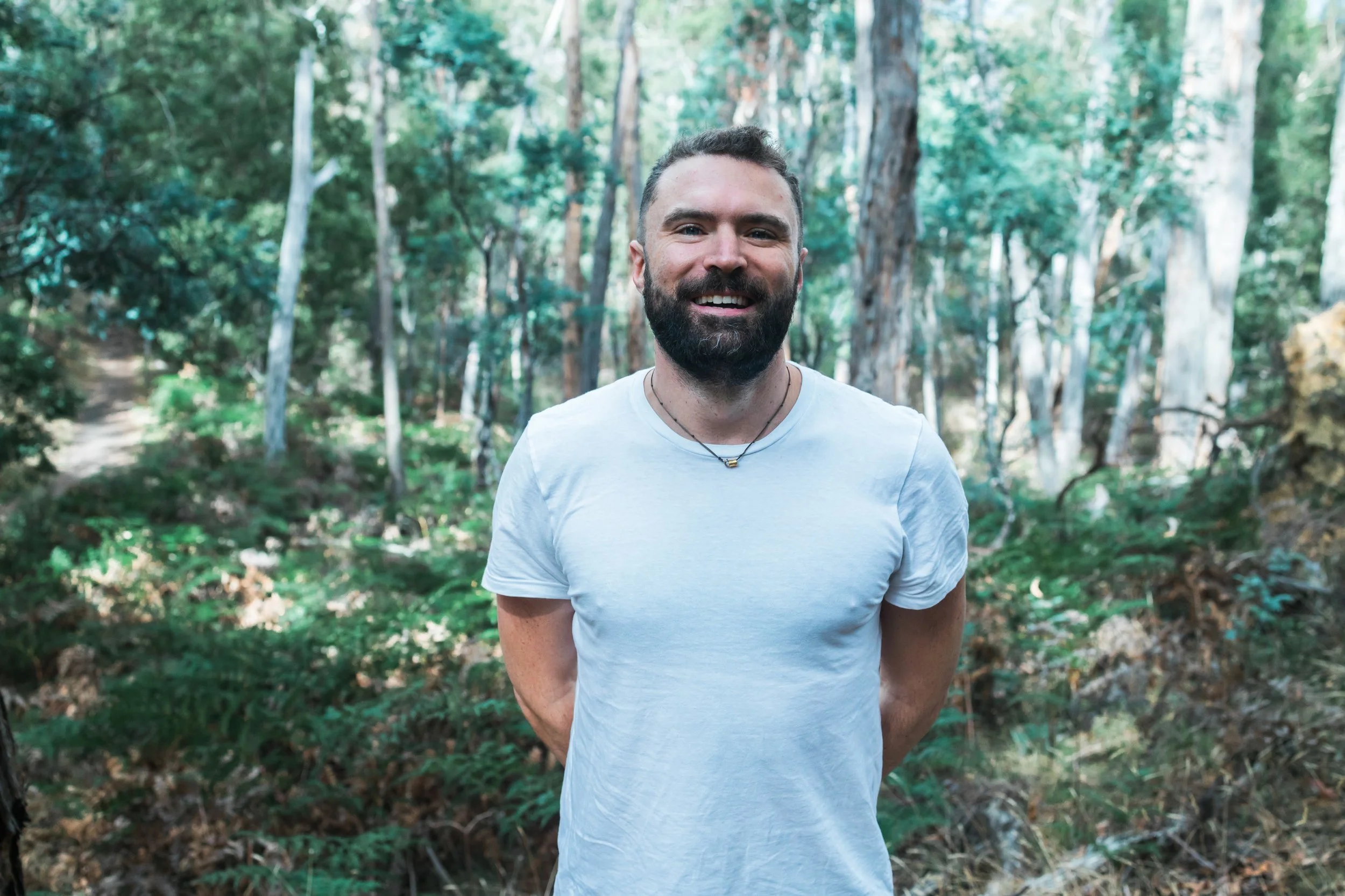 A man with a beard, smiling, wearing a white t-shirt and a necklace, standing with his hands behind his back in a wooded area with trees and greenery.
