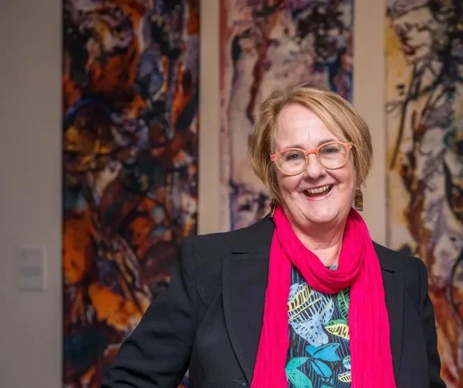 A smiling woman with short blonde hair, glasses, and a red scarf standing in front of colorful abstract artwork.