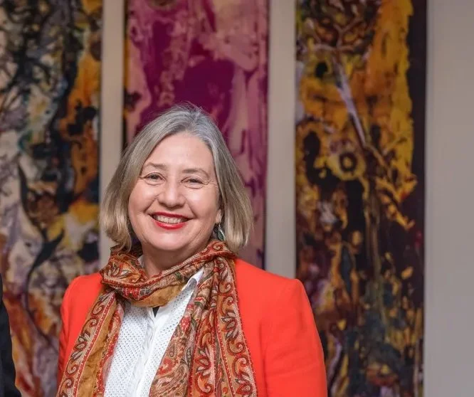 A smiling woman with gray hair wearing a bright orange blazer and patterned scarf, standing in front of abstract artwork with yellow, purple, and black colors.