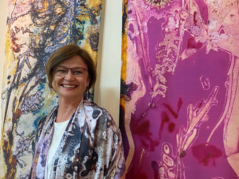 A smiling woman with glasses standing in front of colorful abstract paintings.
