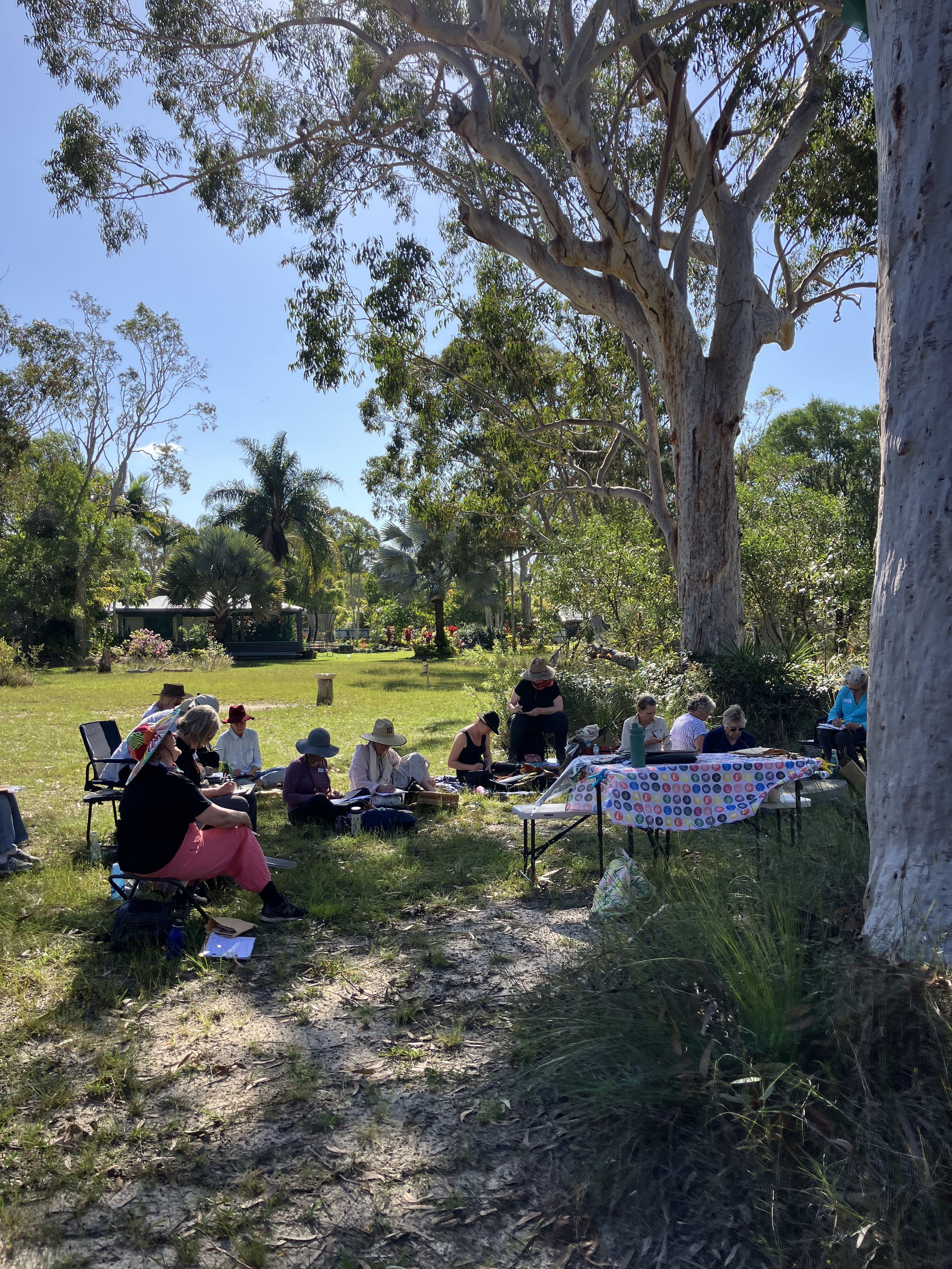 A group of people sitting outdoors under large trees, some taking notes and others reading, with a table covered in a colorful polka dot tablecloth and a lush green park setting in the background.