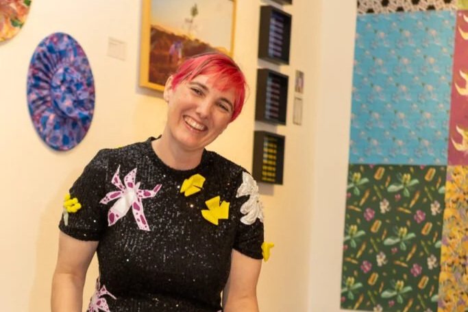 A woman with short pink hair, smiling, wearing a black top decorated with yellow and white fabric cutouts, standing in an art gallery with colorful paintings and artworks on the wall.