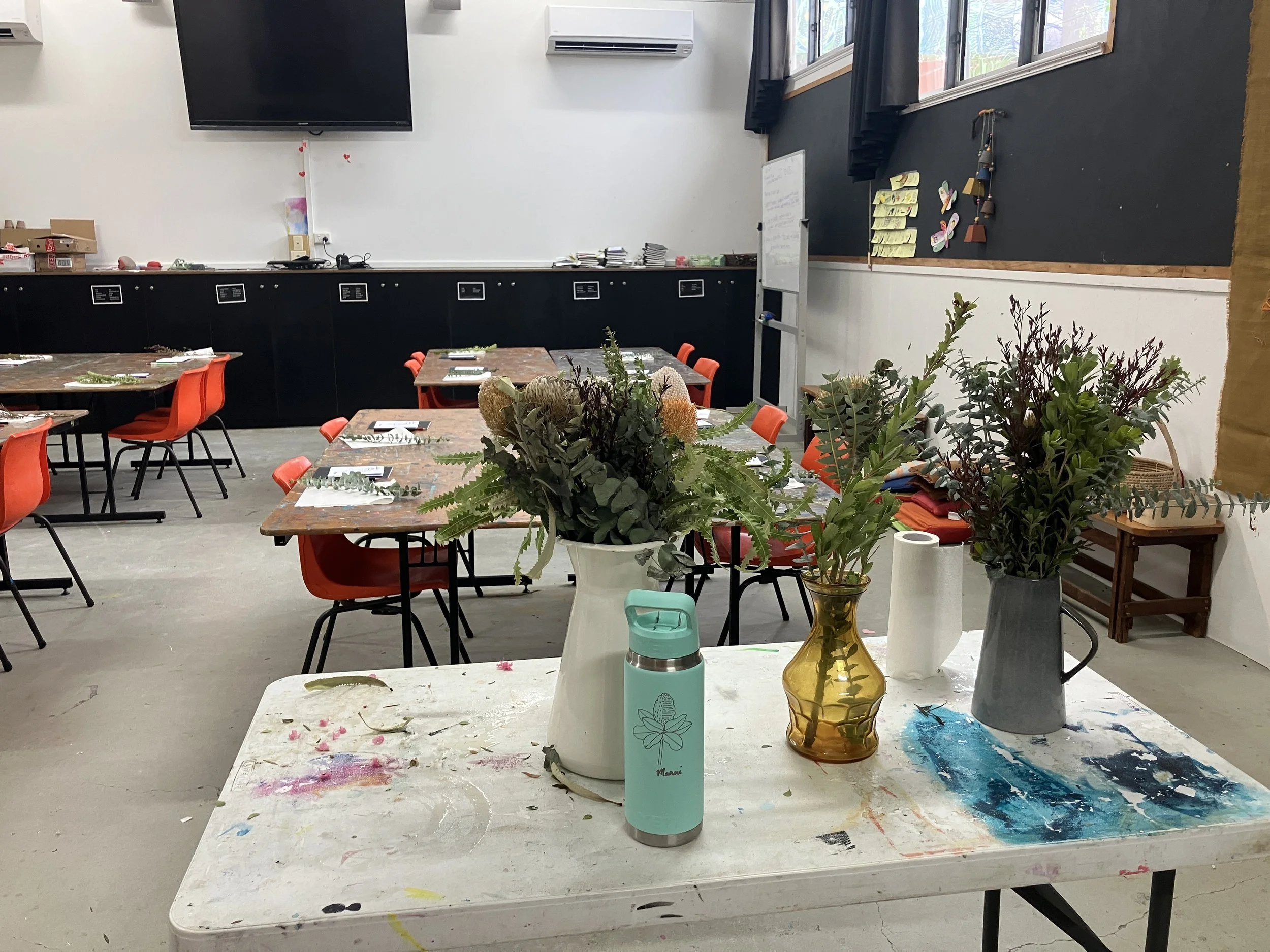 A room decorated for a celebration with tables, chairs, flower vases, and remnants of flowers and paint, suggesting a floral arranging activity or party.