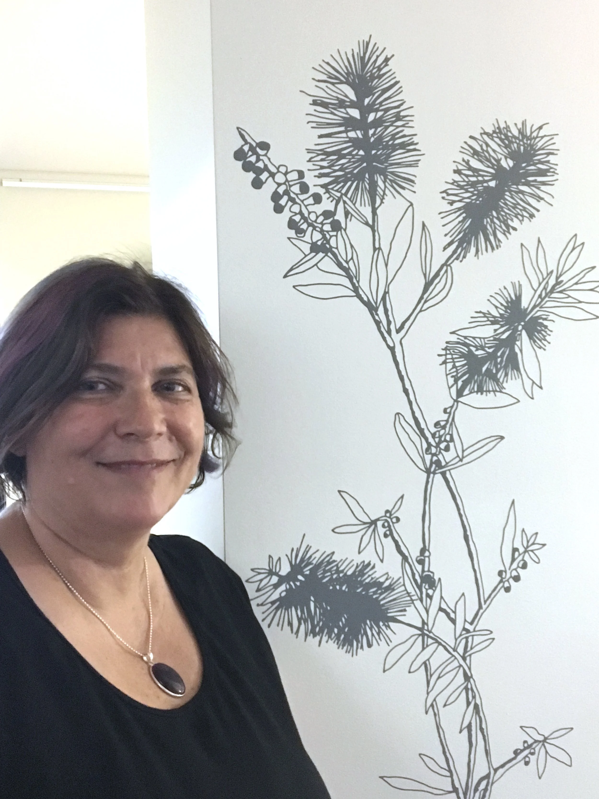 A woman with short hair stands beside a cut out vinyl work on a wall. The work is of Wallum Bottlebrush.