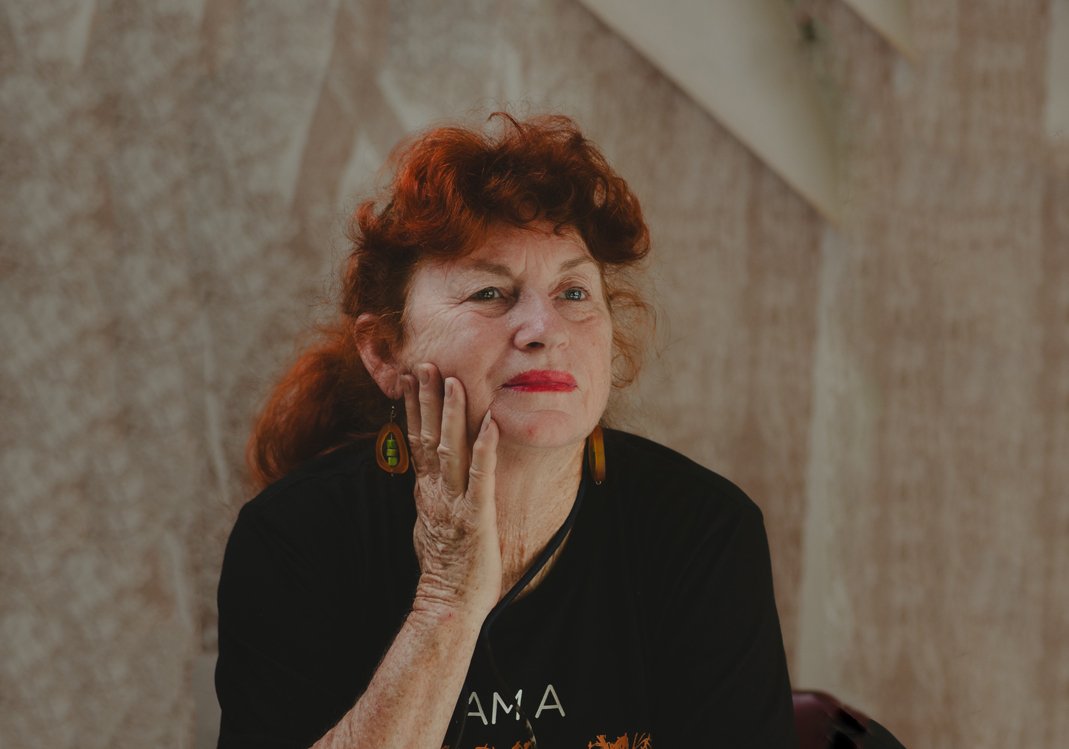 A woman with curly red hair and glasses wearing a colorful red, pink, and purple patterned dress, standing in front of a large artwork featuring detailed botanical illustrations at an art gallery.
