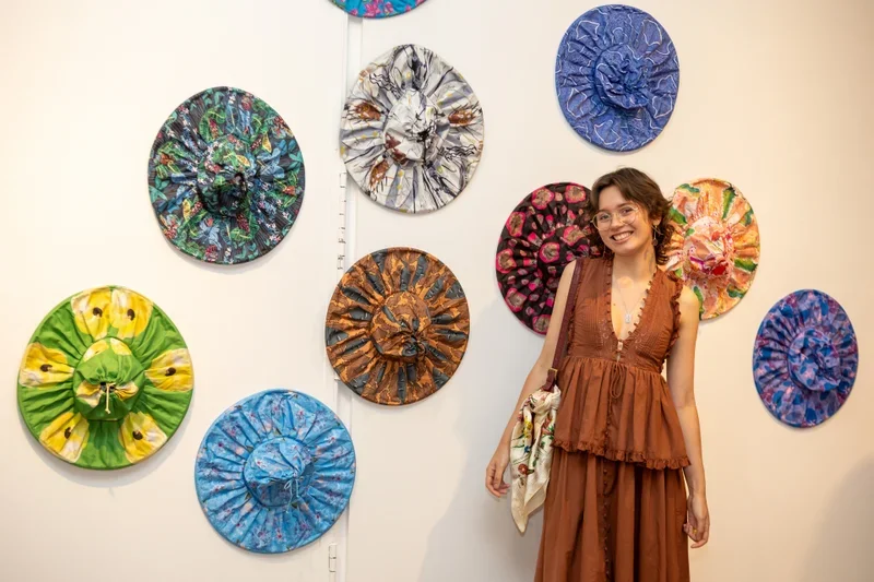 A woman in a brown dress standing next to an art display of colorful, textured hats mounted on a white wall. The hats are arranged in a circular pattern and each has a unique design and color scheme.