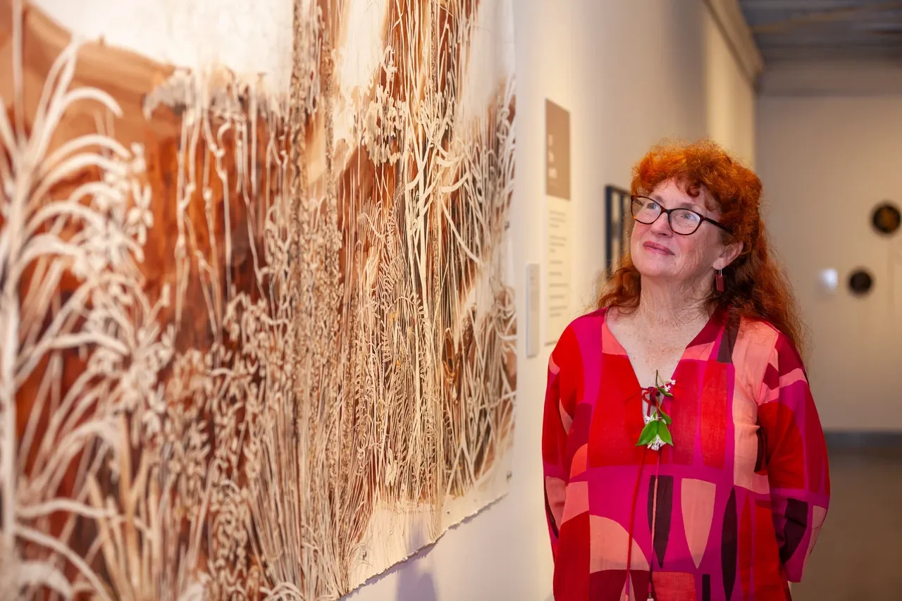 A woman with curly red hair and glasses wearing a colorful red, pink, and purple patterned dress, standing in front of a large artwork featuring detailed botanical illustrations at an art gallery.