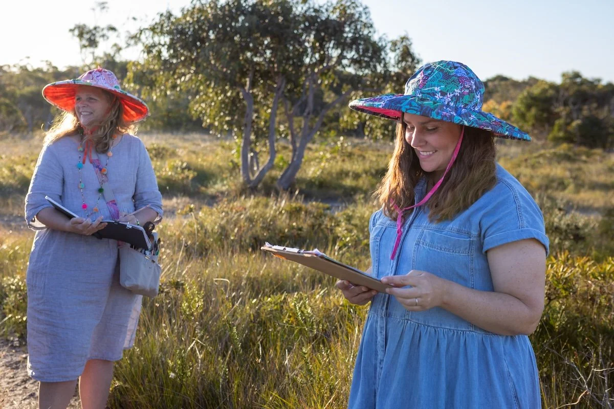 Two women standing outdoors in a grassy area, wearing colorful wide-brimmed hats, smiling and looking at clipboards.