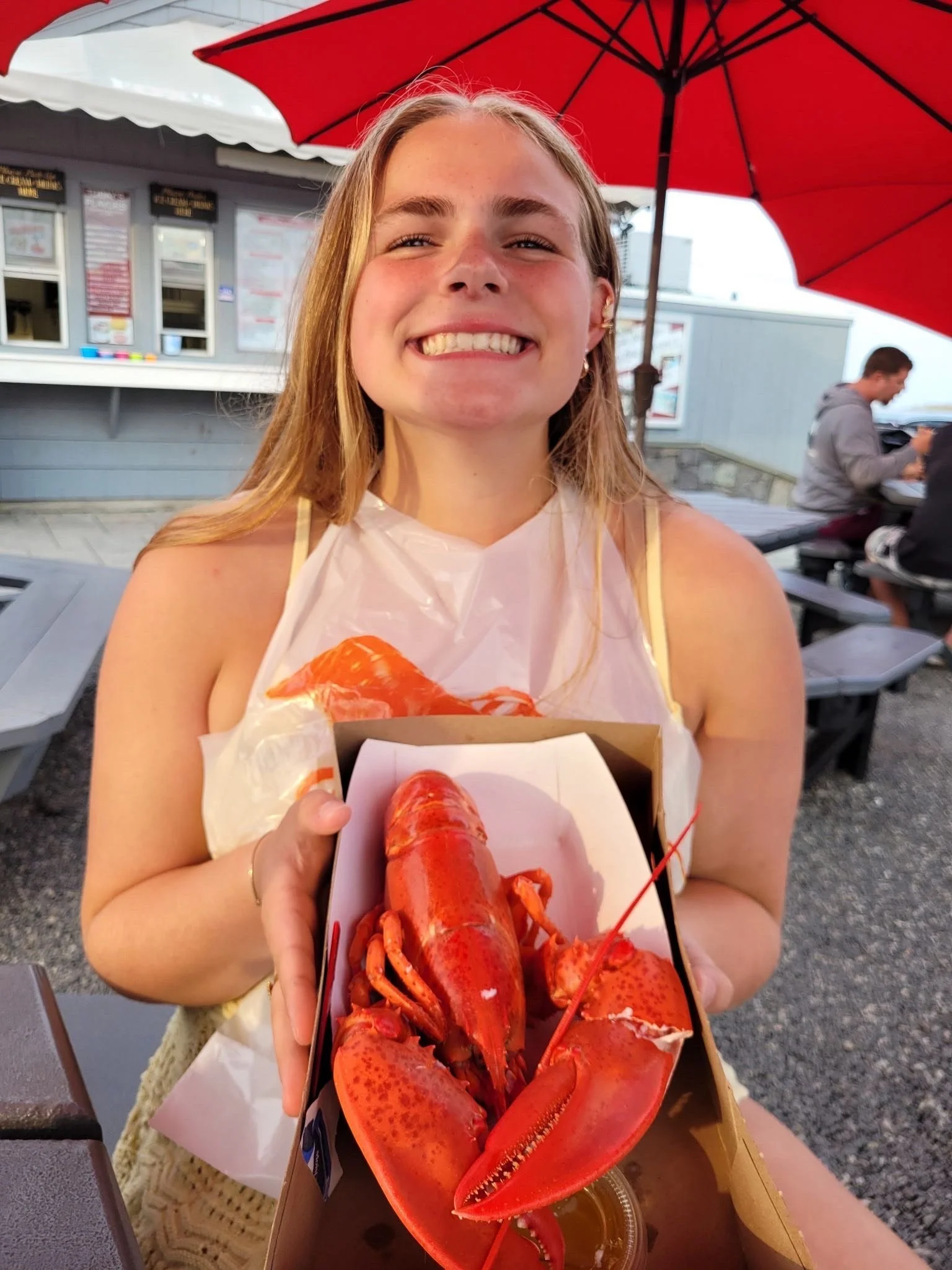 A smiling young woman holding a box with a cooked lobster, sitting outdoors at a restaurant with a red umbrella.