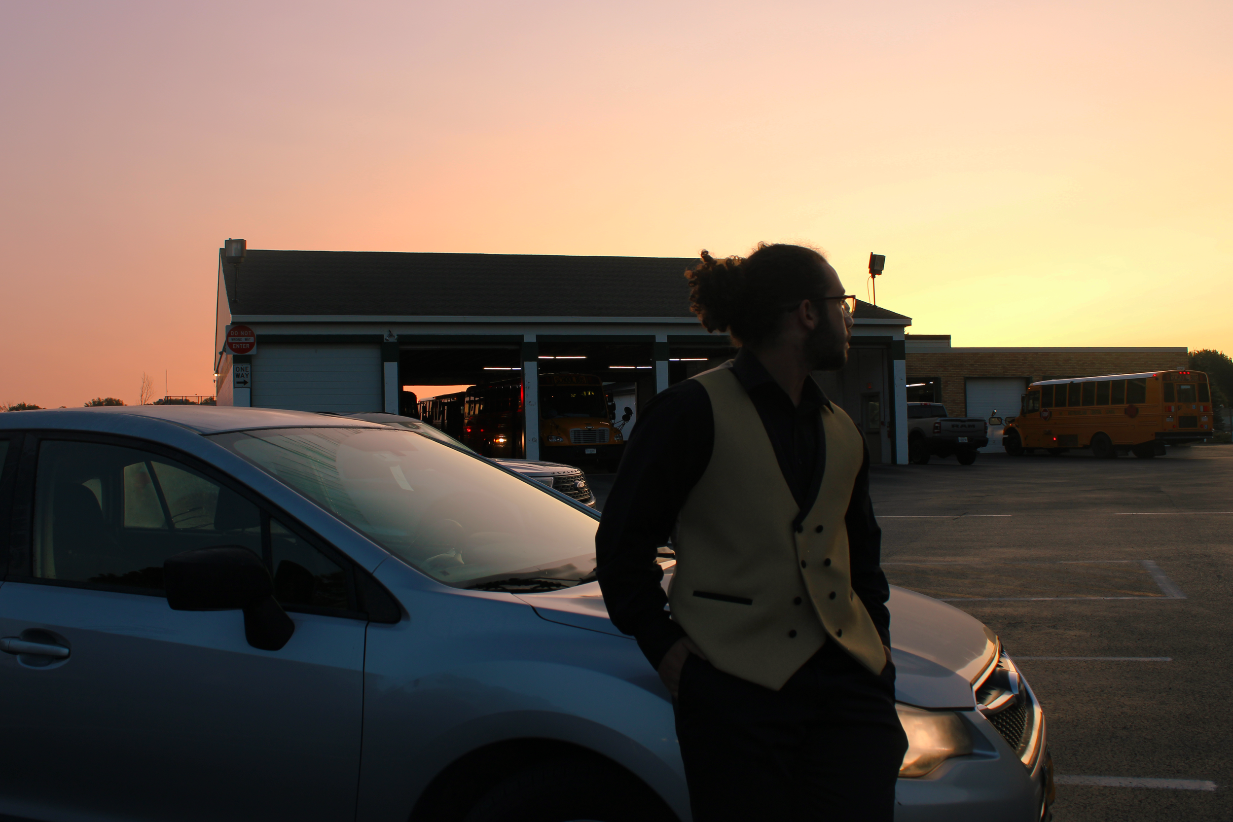 A man with glasses and a beard, dressed in a black shirt and beige vest, stands outside with his hands in pockets, leaning against a blue car, at sunset in a parking lot with school buses and a building in the background.