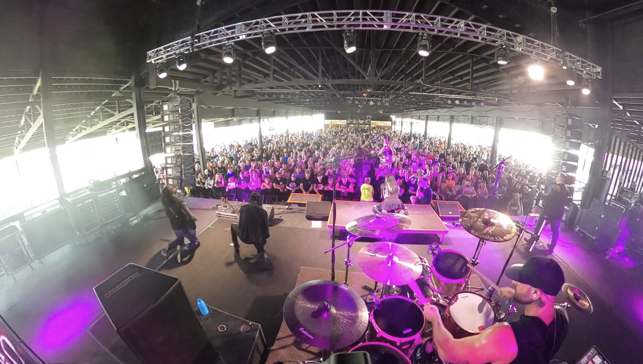 View from a stage looking out towards a large crowd of people at a concert or music event inside a covered venue with stage equipment, drum set, and musicians.