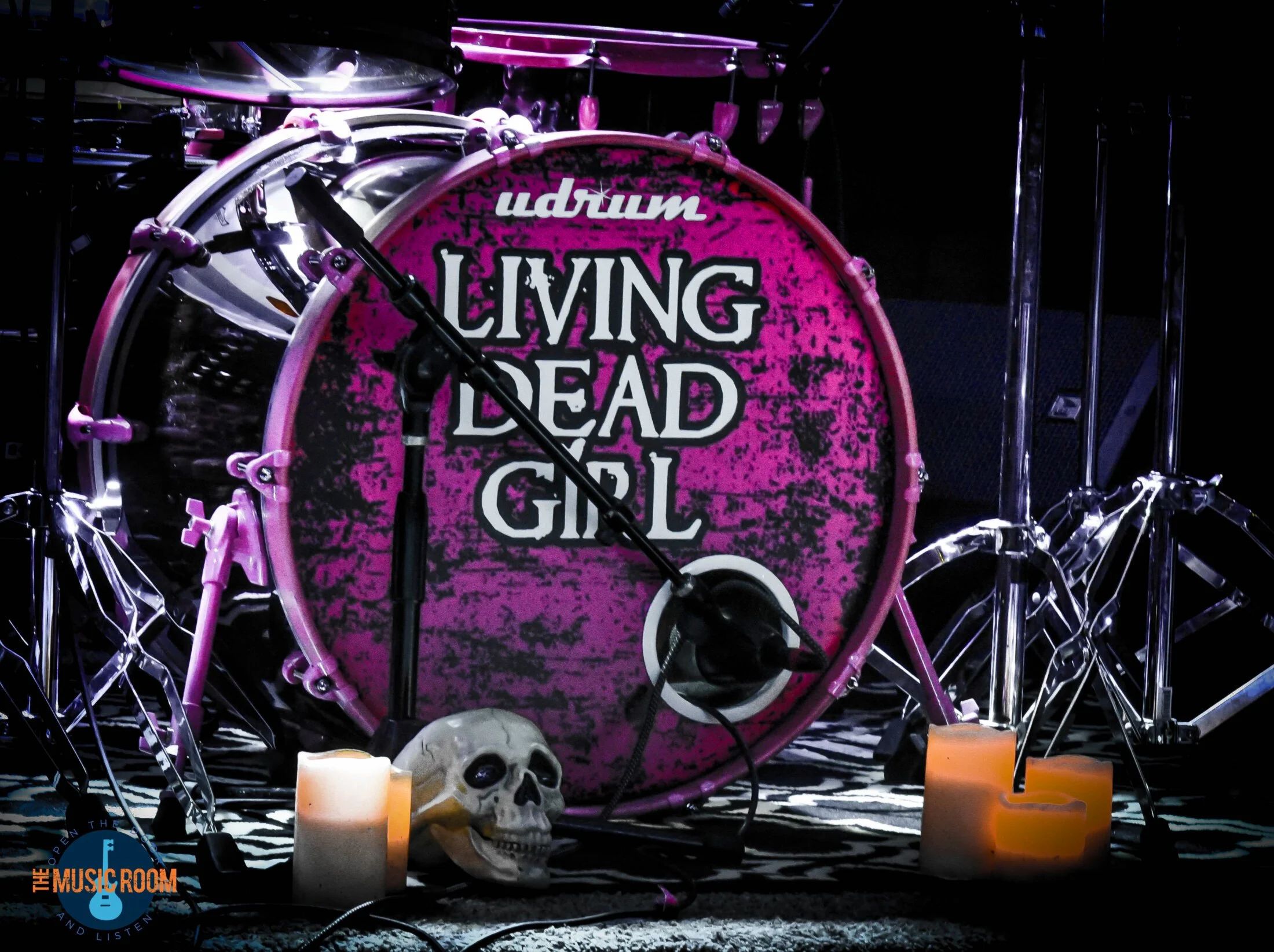 A pink drum set with the words 'drum living dead girl' on the bass drum, surrounded by candles, a skull, and musical equipment, set on a black-and-white patterned floor.