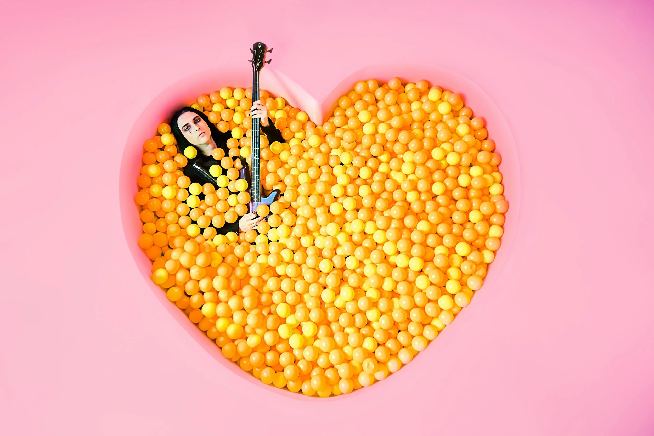 A woman lying in a pink heart-shaped ball pit filled with yellow and orange balls, holding a guitar, against a pink background.
