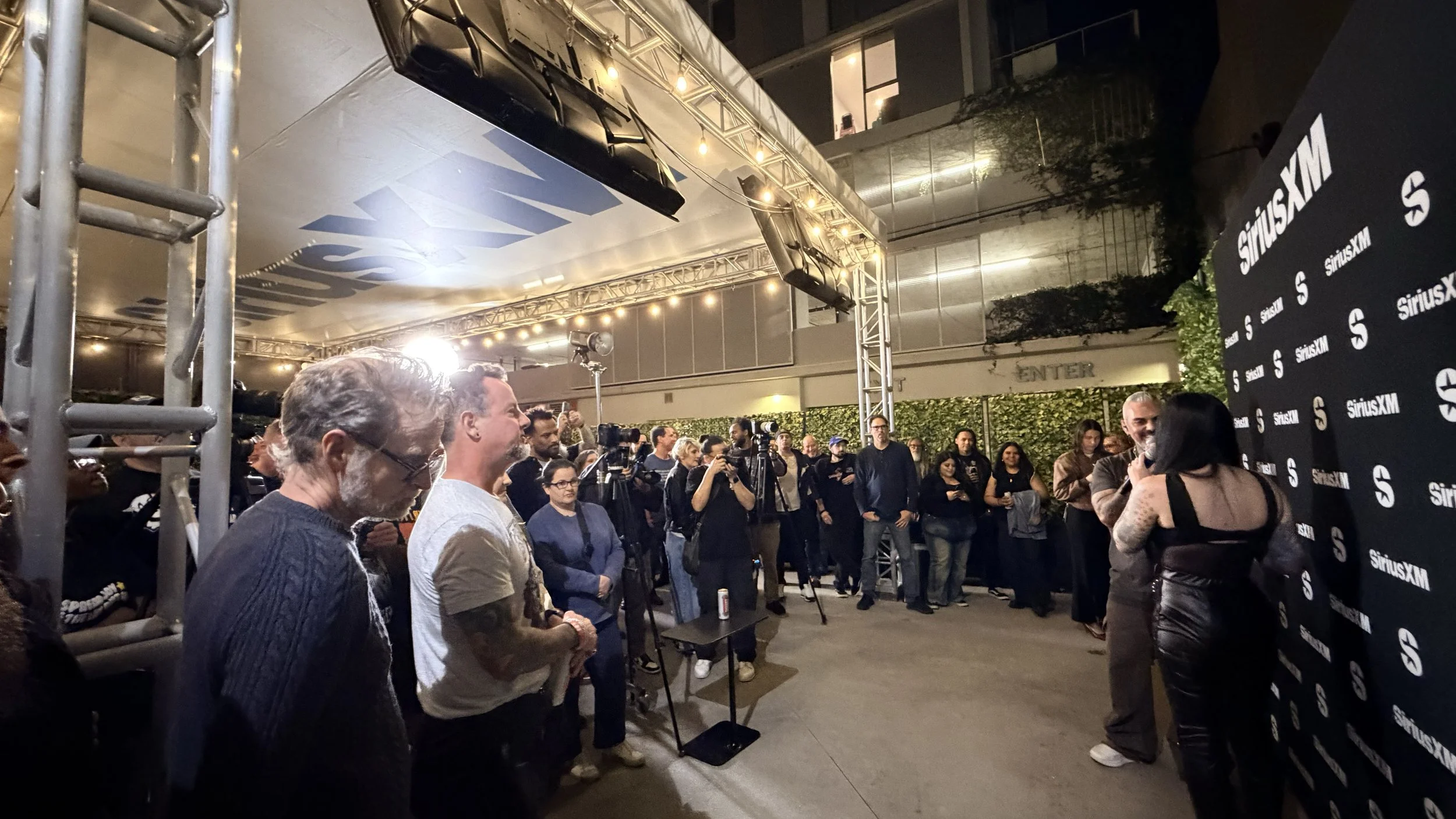 People gathered outdoors at night for a SiriusXM event, with a woman being interviewed near a Black SiriusXM branded backdrop and photographers capturing the moment.