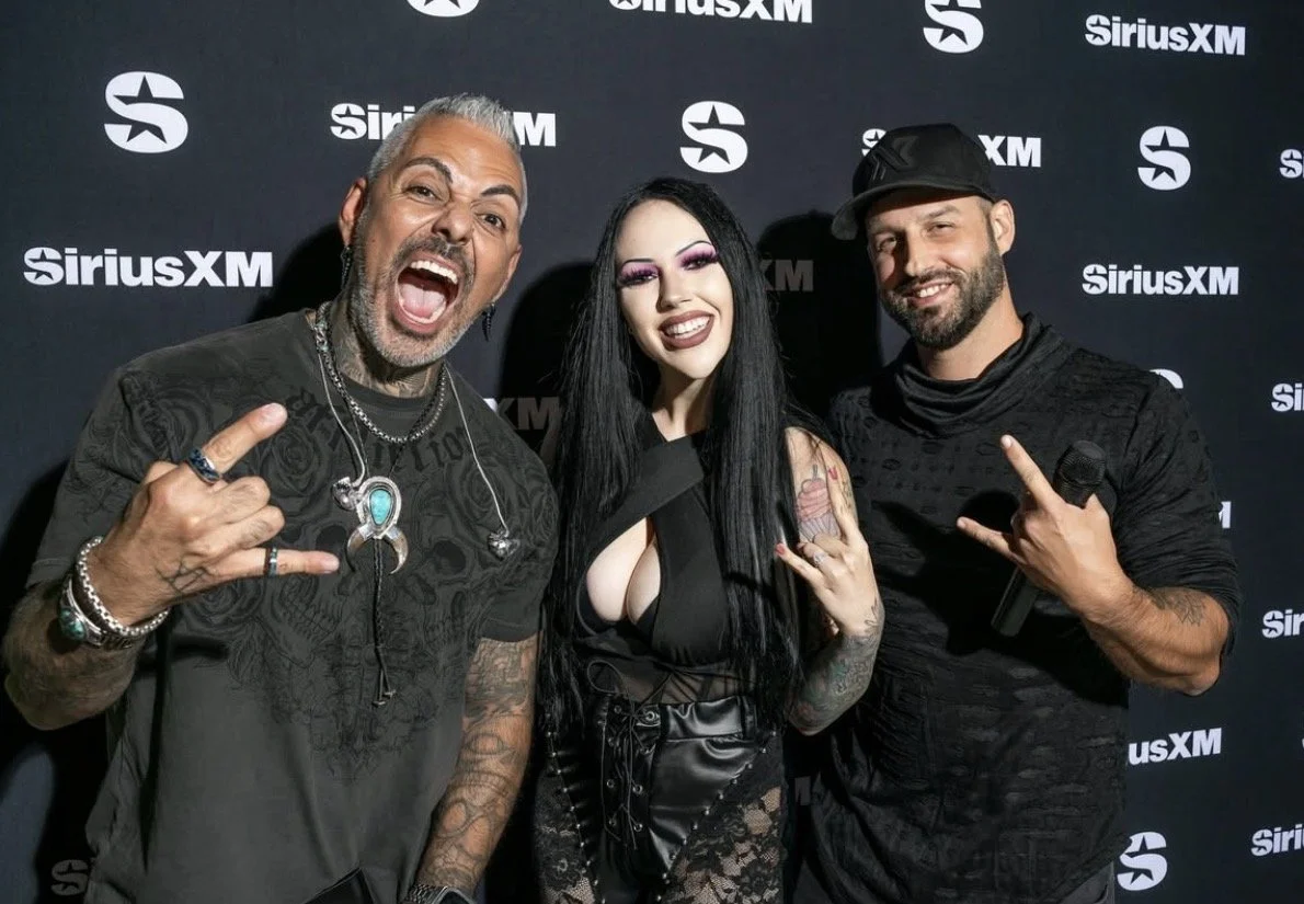 Three people at a SiriusXM event posing for a photo, with a dark backdrop featuring SiriusXM logos. The person on the left is a man with short gray hair, tattoos, and singing or shouting with his mouth open. The person in the middle is a woman with long black hair, pronounced makeup, wearing a revealing black outfit, and smiling. The person on the right is a man with a beard, wearing a black cap and shirt, and holding a microphone while making a hand gesture.