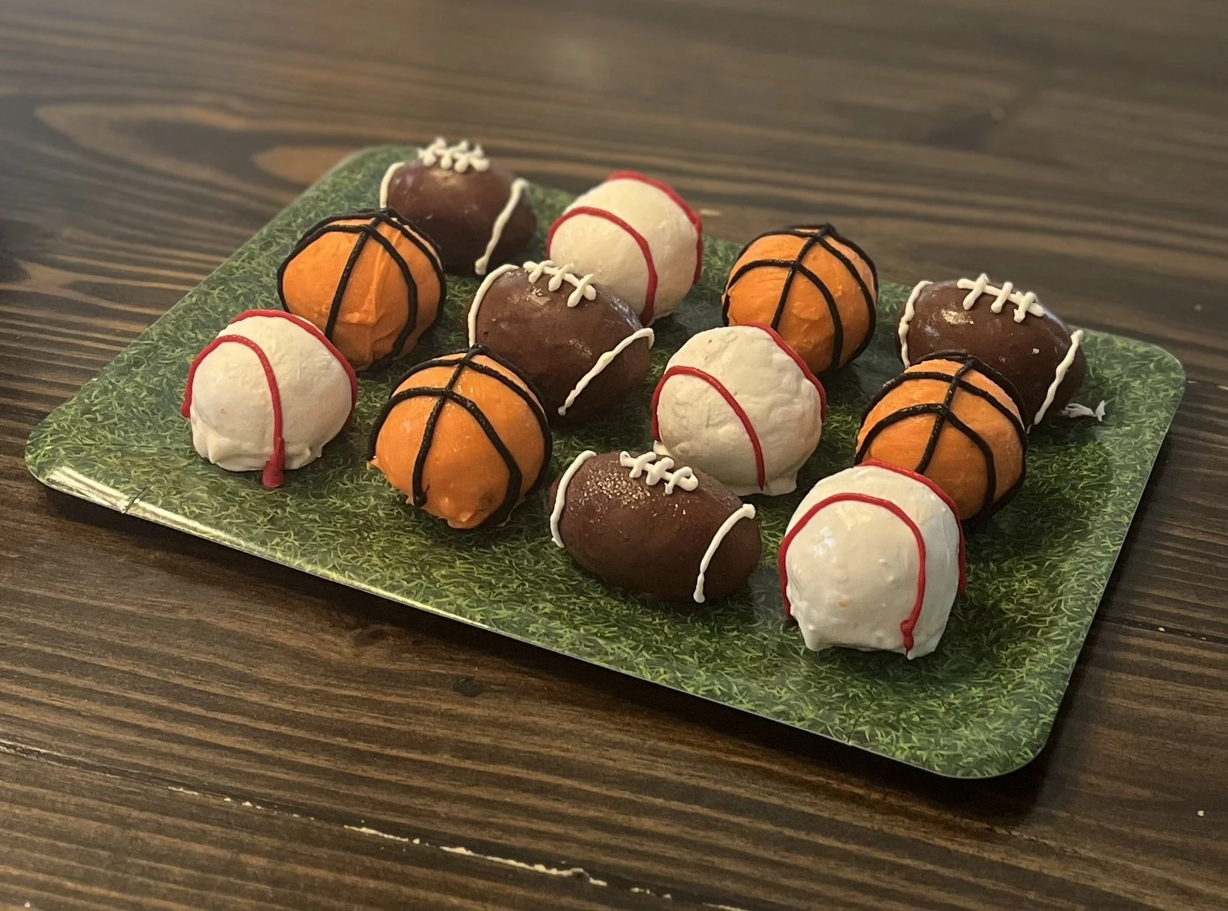Plate of basketball-themed cake pops decorated as basketballs and basketball jerseys, placed on a green patterned platter on a wooden table.