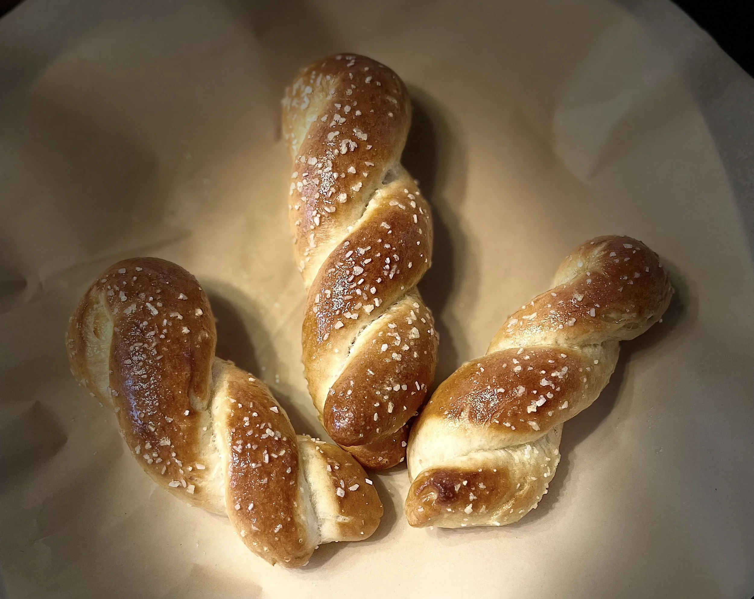 Three twisted pretzels with coarse salt on top, inside a paper baking sleeve.