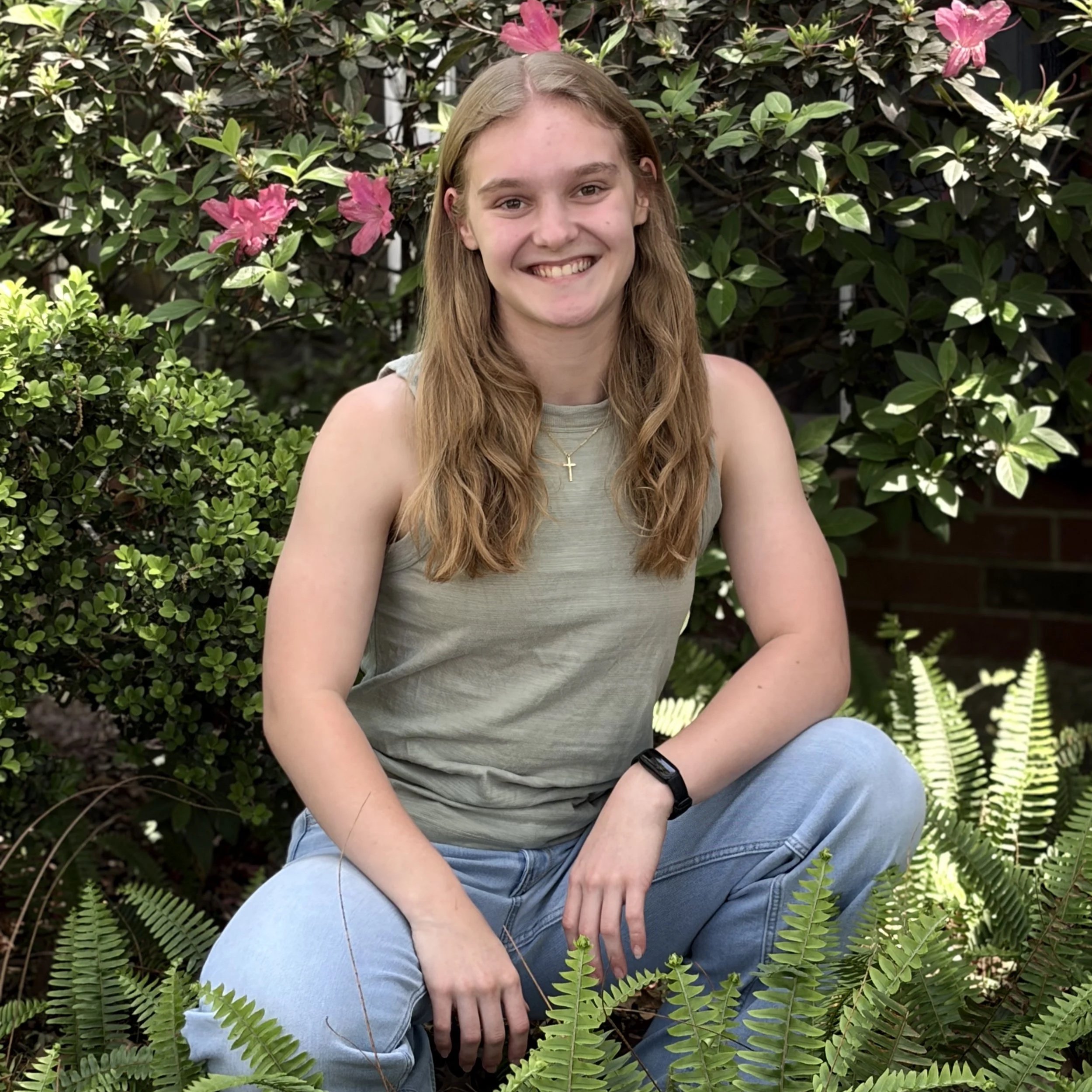 A young woman with long wavy blonde hair smiling, sitting outdoors amid greenery and pink flowers.