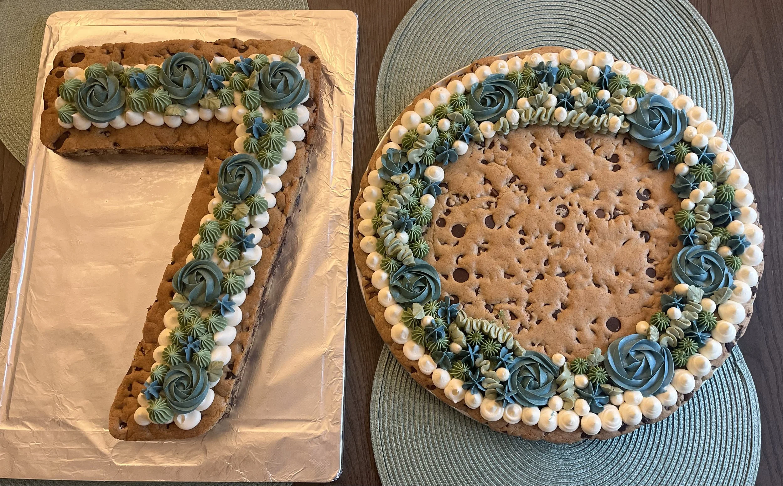 A cake shaped like the number seven with chocolate chips inside, decorated with green and white frosting flowers and pearls, and another round cookie cake decorated similarly with frosting flowers along the top edge.