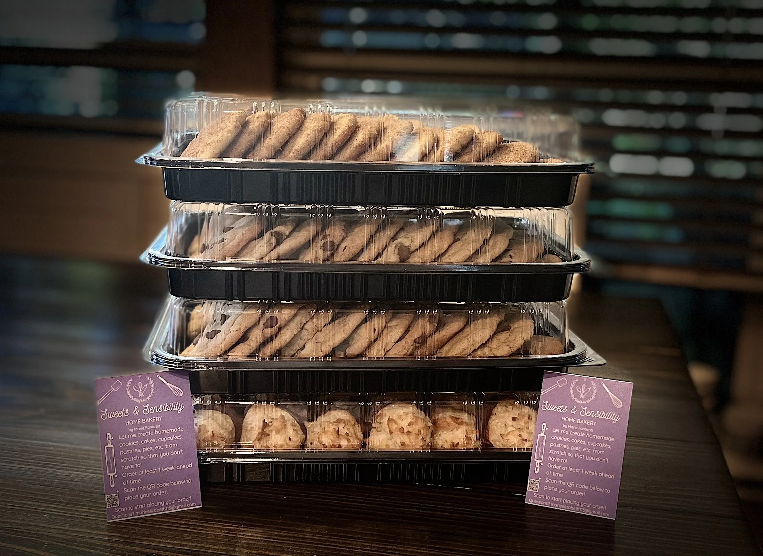 Stacked clear plastic food containers filled with cookies and cookies with white chocolate chips, placed on a wooden surface.