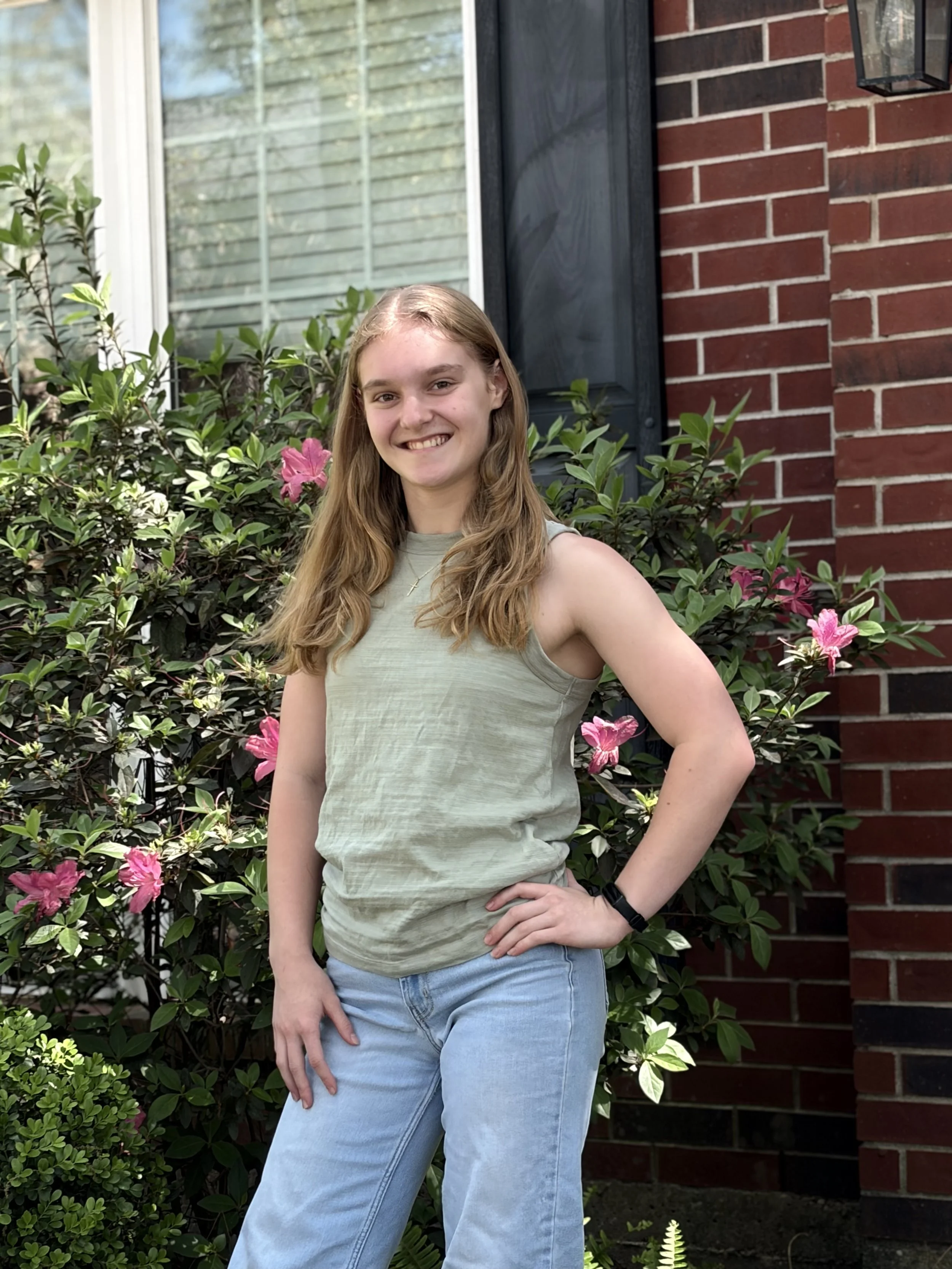 A young woman with long, wavy blonde hair smiling and posing outdoors in front of greenery and a brick house with a window and black door.