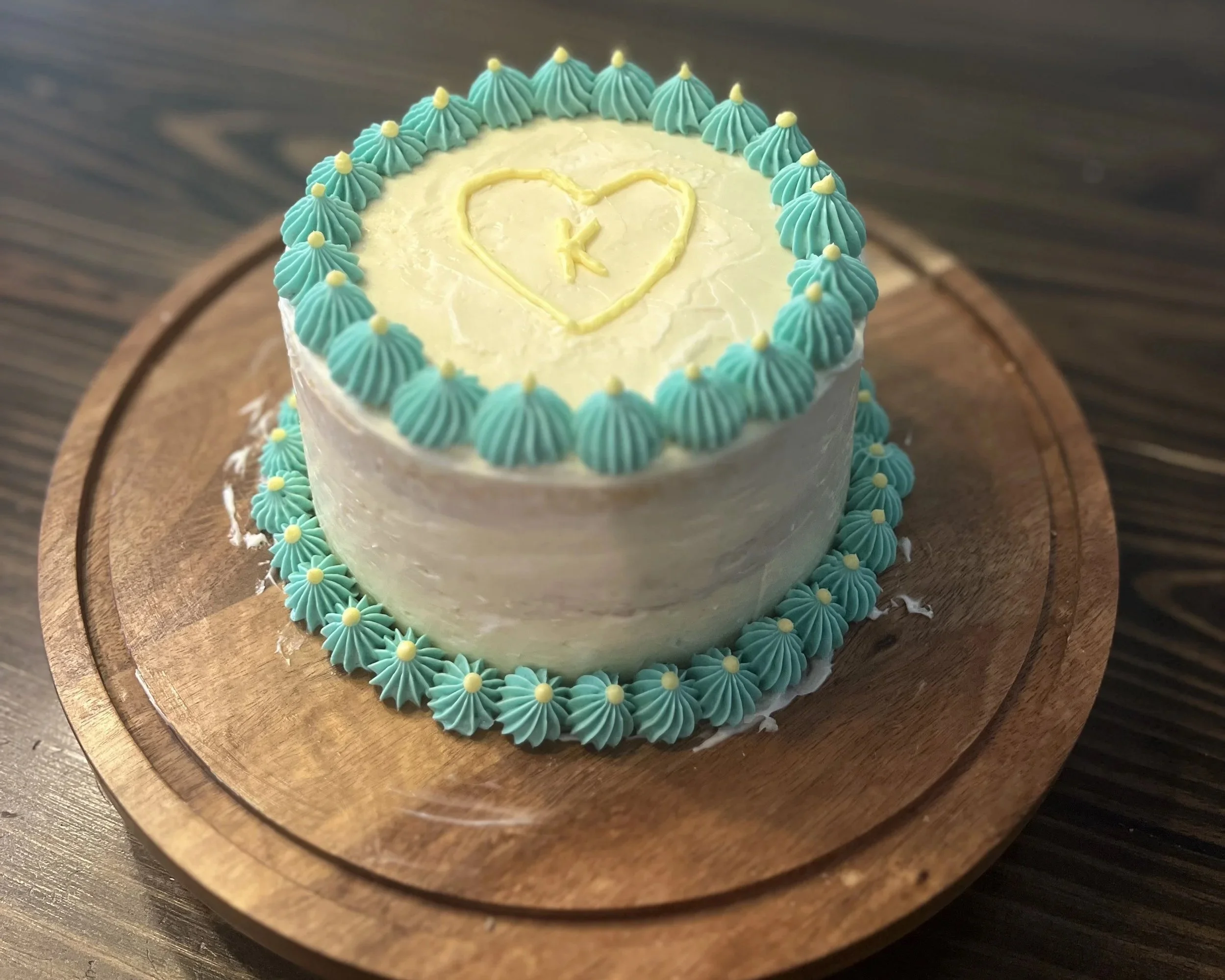 A round cake decorated with light blue piping around the top edge and bottom edge, featuring small yellow dots on the decorations. The cake has a white frosting exterior with a yellow heart shape outlined on the top, containing the letter 'K' inside.