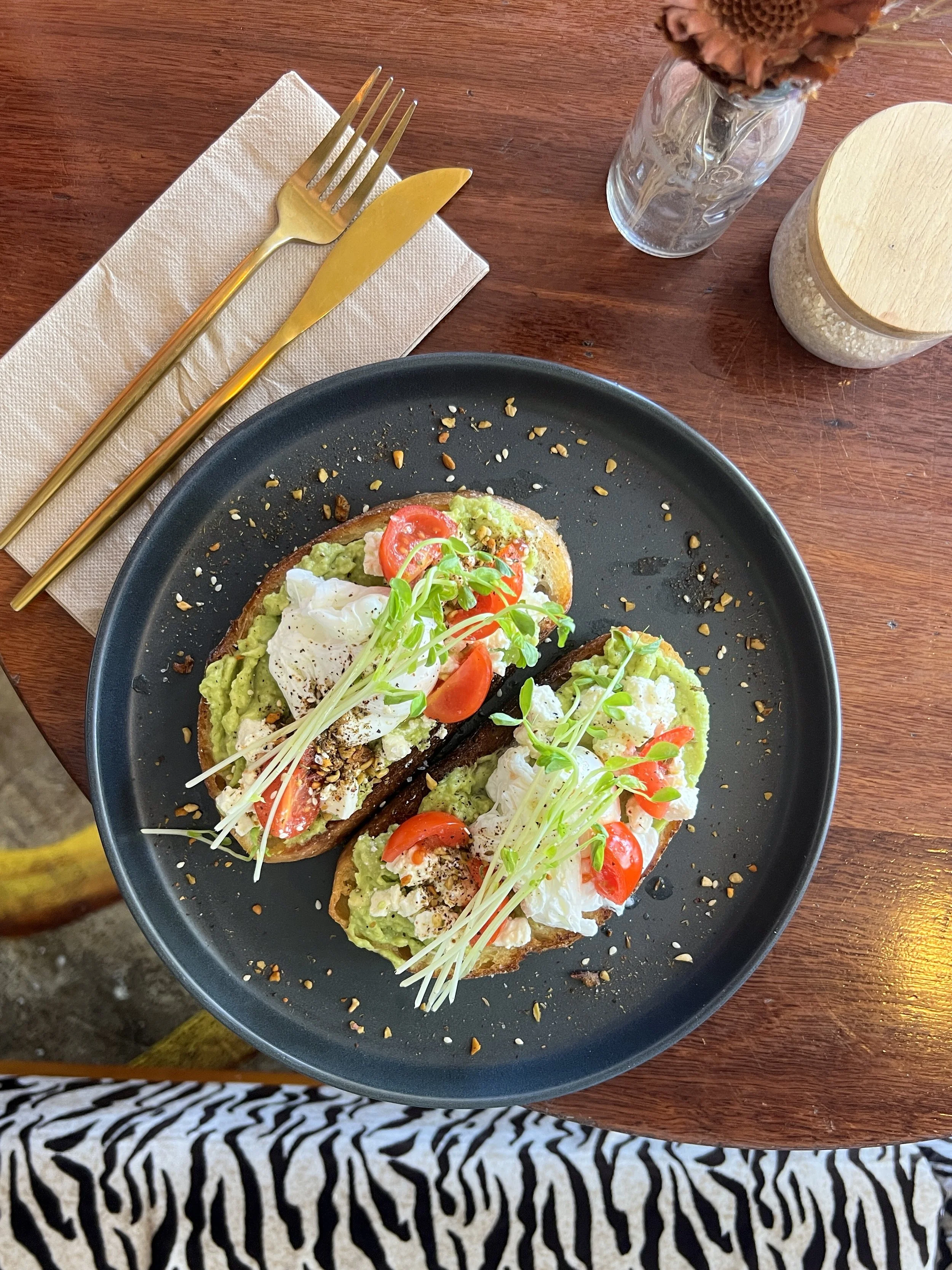 Two avocado toast slices topped with poached eggs, cherry tomatoes, and microgreens on a black plate.