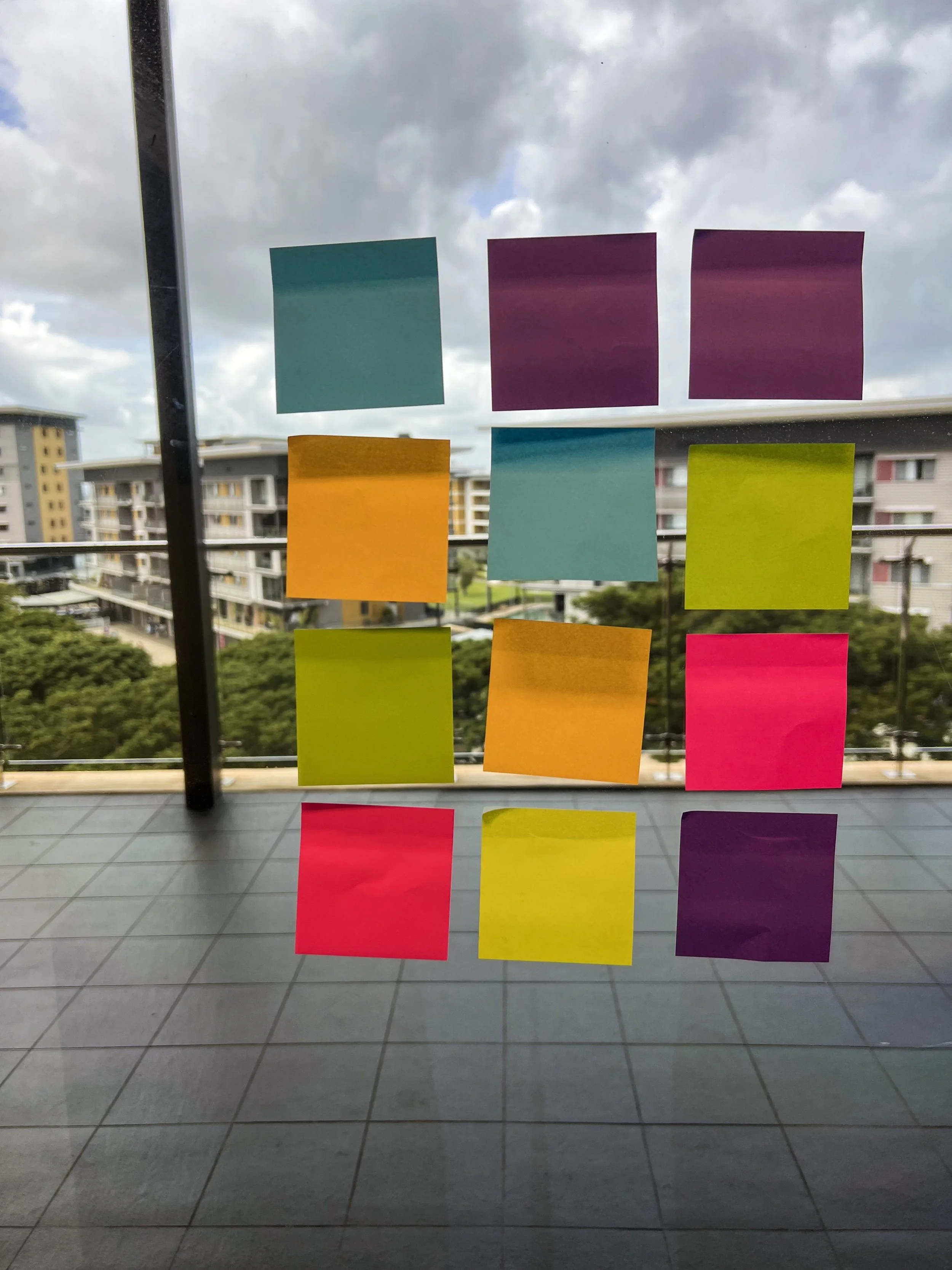 Multiple colorful sticky notes adhered to a glass window, with a background of a cloudy sky, residential buildings, and trees.