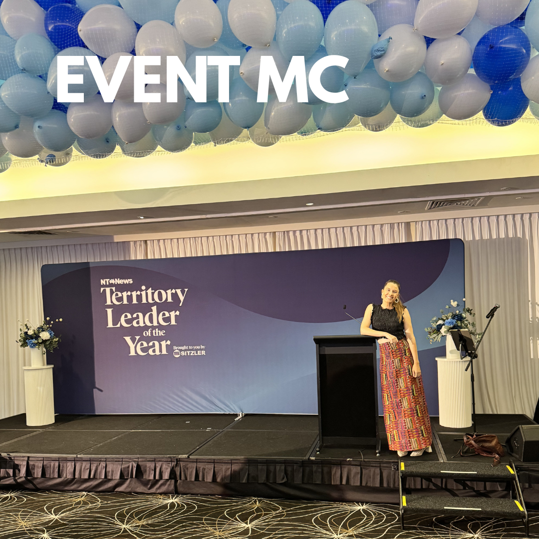 A woman standing on a stage with a podium and microphone, smiling. Behind her is a backdrop with the words 'NT News Territory Leader of the Year' and flowers on stands. Overhead, white and blue balloons are arranged in a cluster.