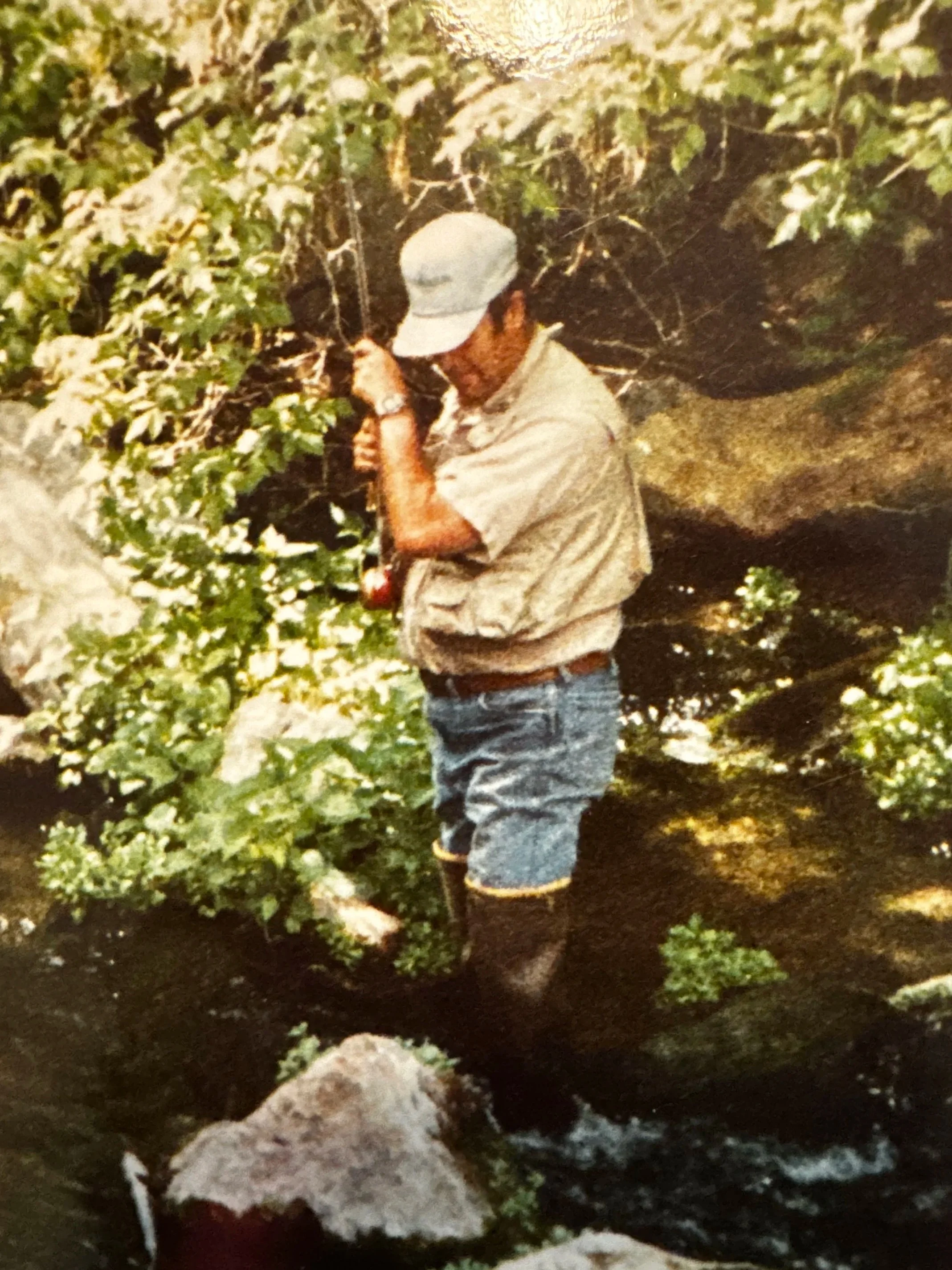 Man standing outdoors among greenery, wearing a light-colored cap, beige shirt, and blue jeans, with a rock and soil ground.
