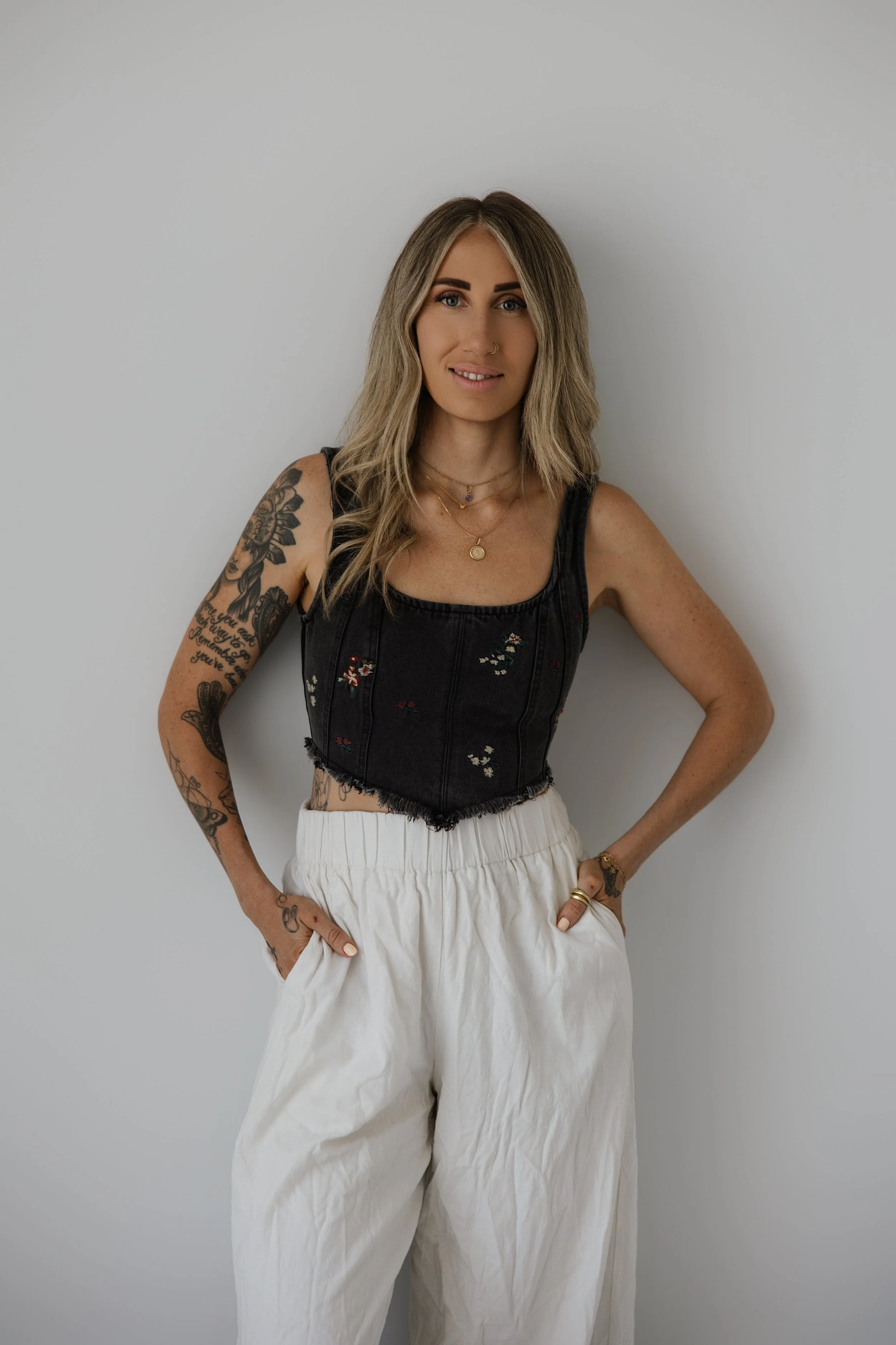 A woman standing against a plain light gray wall, wearing a black sleeveless top with embroidery, white high-waisted wide-leg pants, and gold jewelry, with tattoos on her arms and a nose ring.
