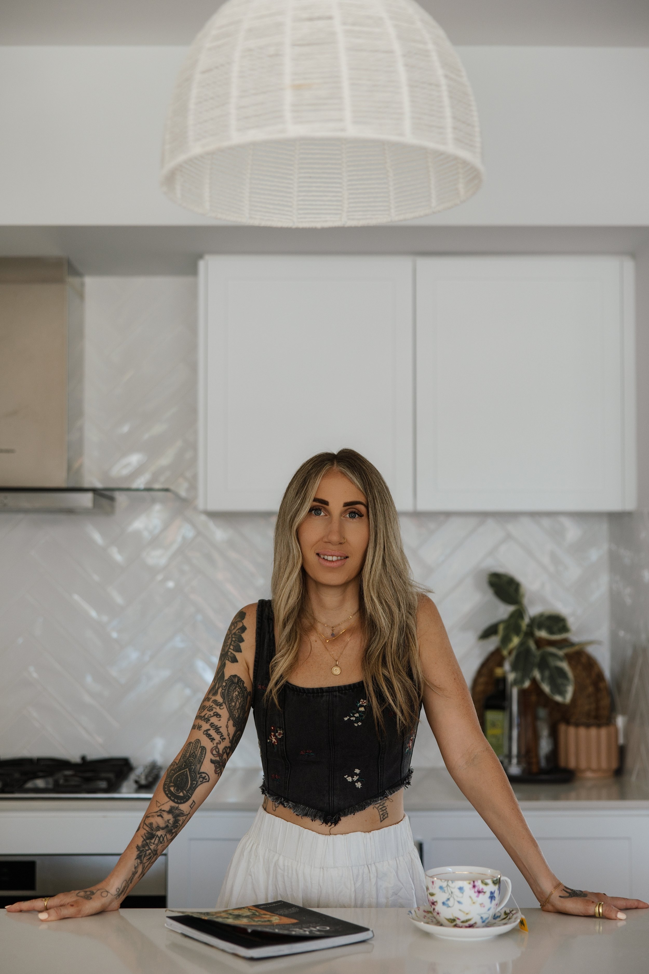A woman with long blonde hair and tattoos on her arms is standing in a modern kitchen, leaning forward with her hands on the counter. She is wearing a black sleeveless top, white pants, and gold jewelry. On the counter in front of her are a magazine and a teacup with a floral pattern.