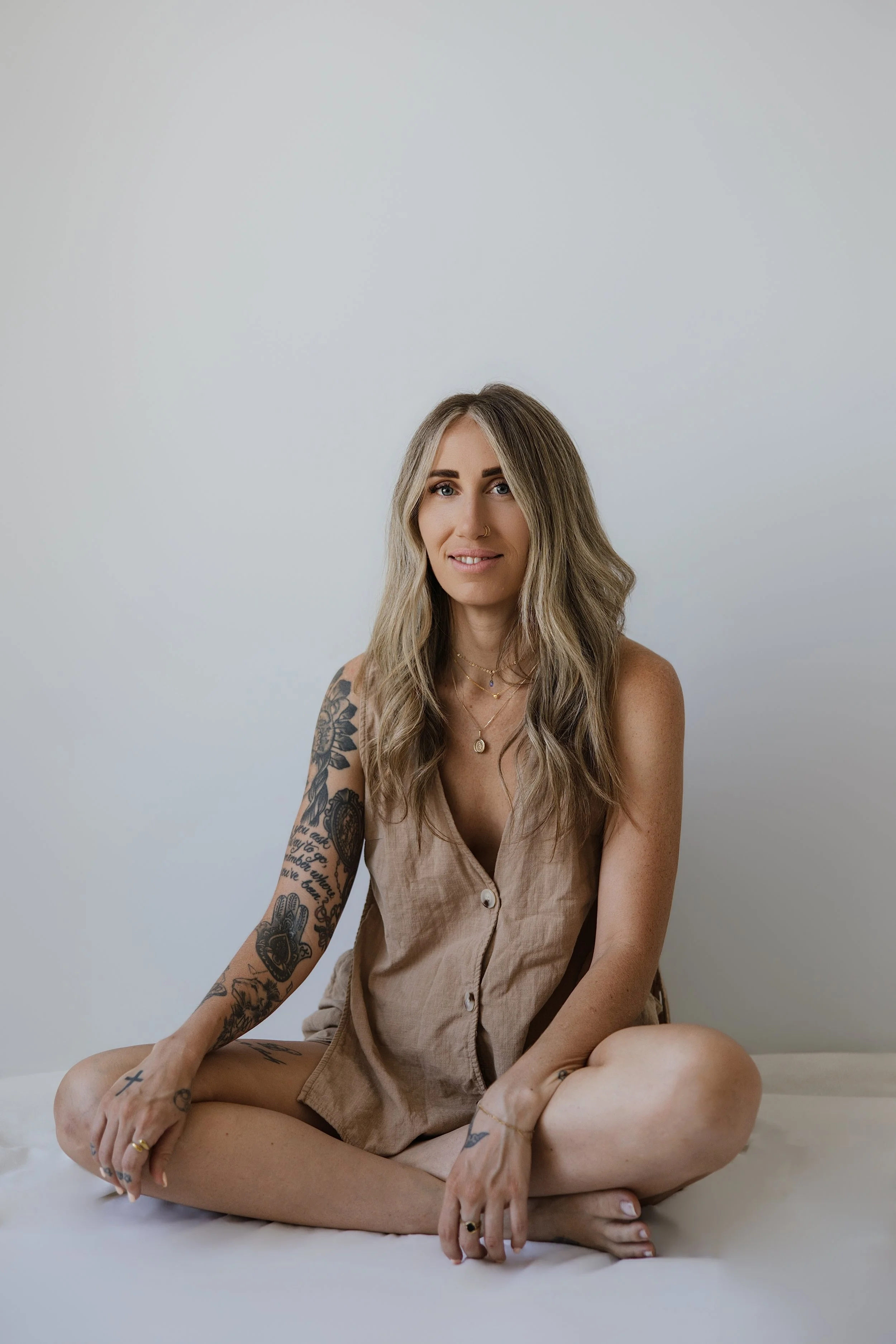 A woman with long, wavy blonde hair, tattoos on her left arm, wearing a beige sleeveless button-down dress, sitting cross-legged on the floor against a plain light-colored wall.