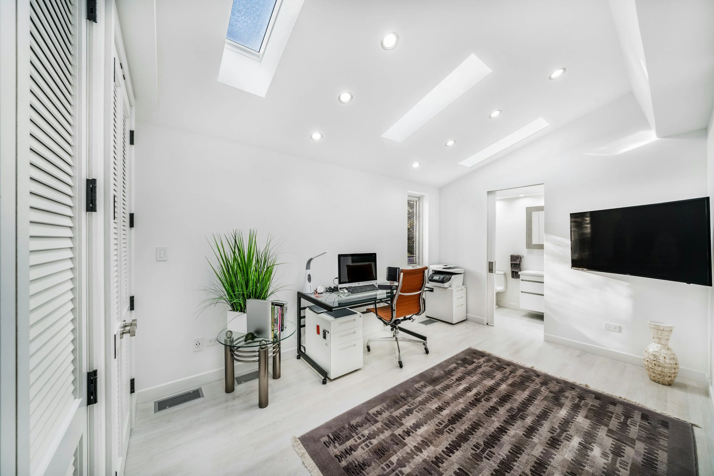 A modern white office with a skylight, ceiling lights, and a window. There is a desk with a computer, a chair, a printer, and some office supplies. A green plant sits on a small table. A patterned rug covers part of the light-colored floor.