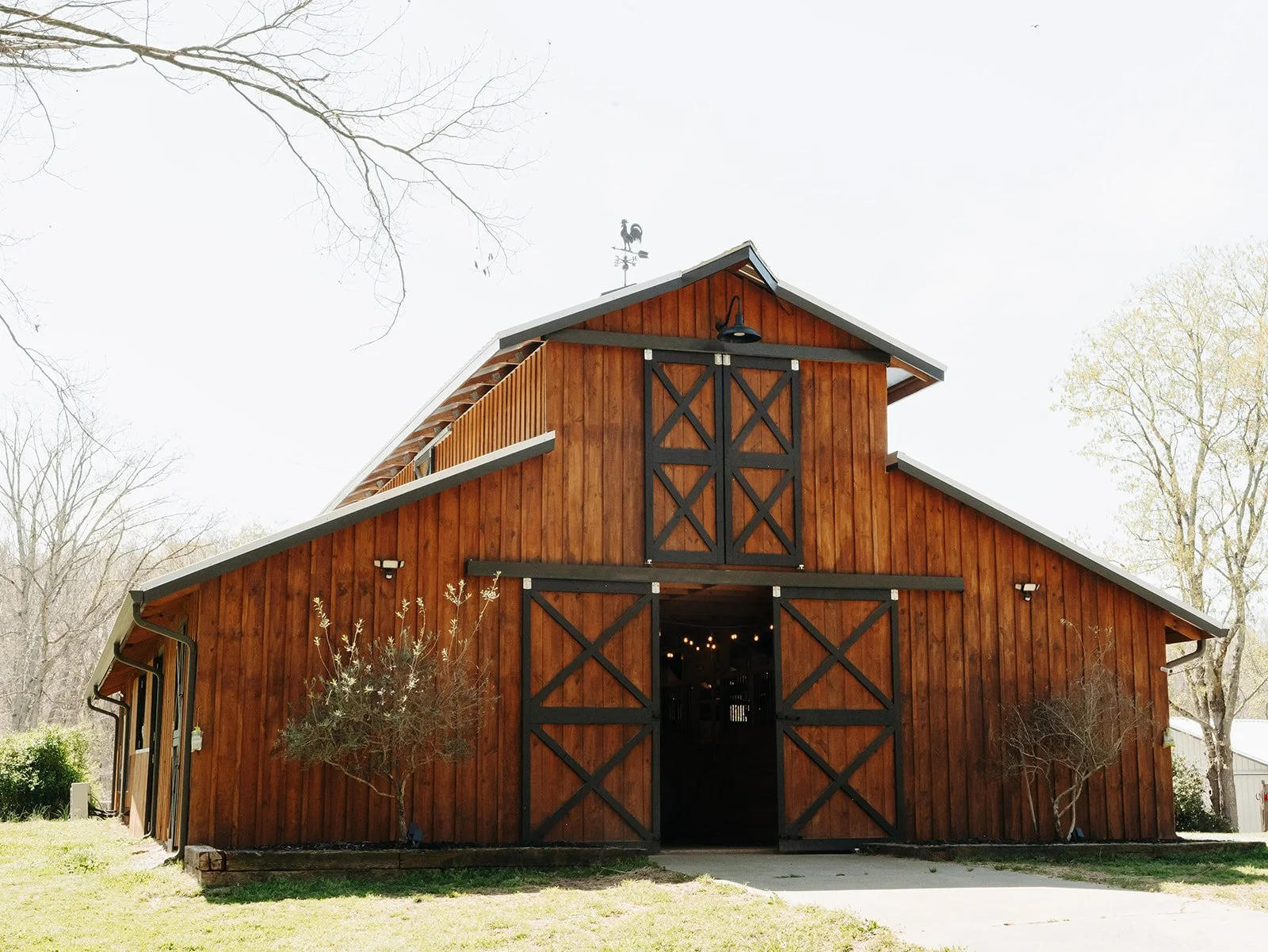 modern rustic wood barn with black trim and sliding doors, trees on grass lawn, and a weather vane on roof barn in field