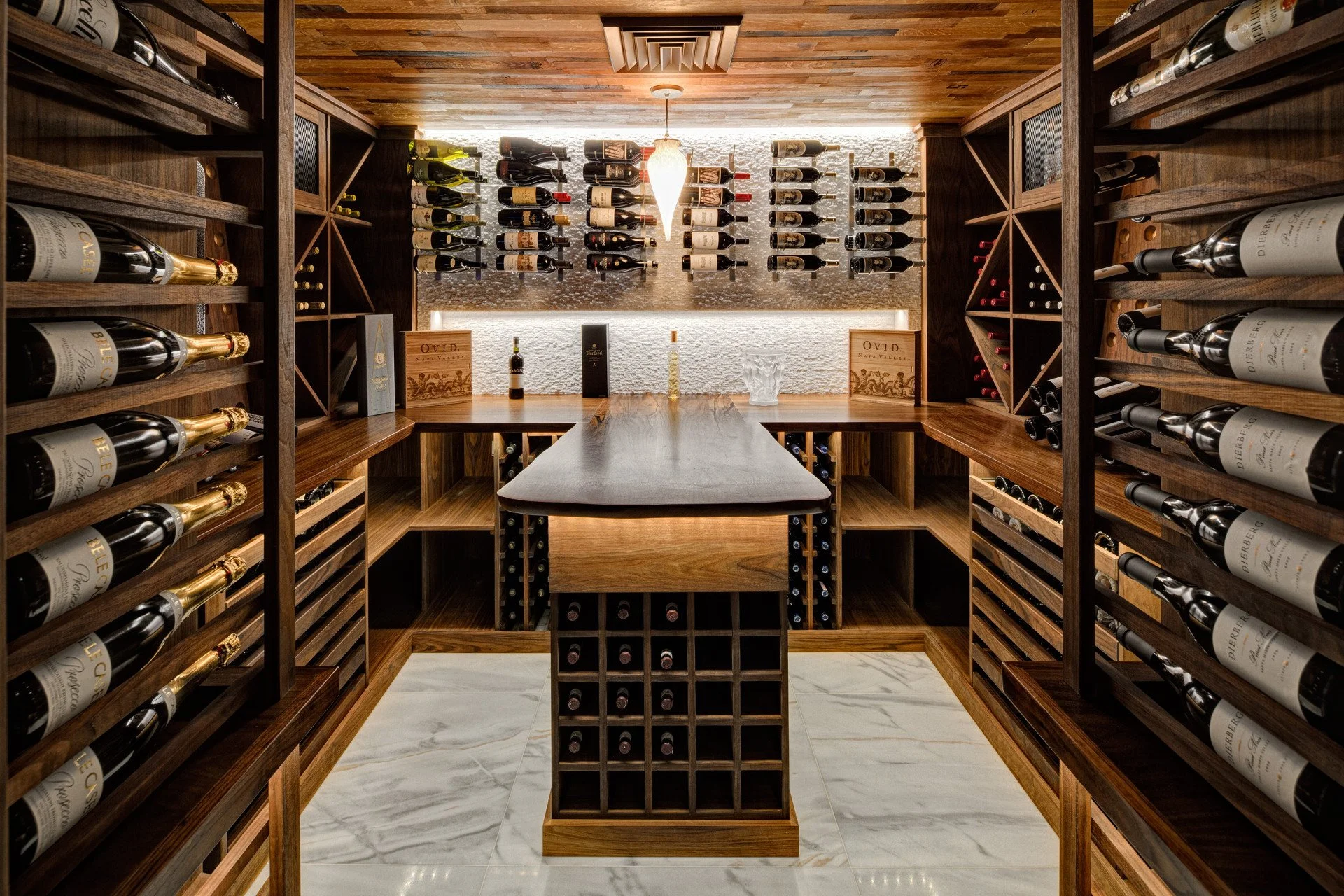 A luxurious wine cellar with wooden shelves filled with wine bottles, a central wooden wine rack, and a small table with bottles on top. The ceiling has a wooden finish with a hanging light fixture, and the back wall features a decorative patch of stone, illuminated by indirect lighting.