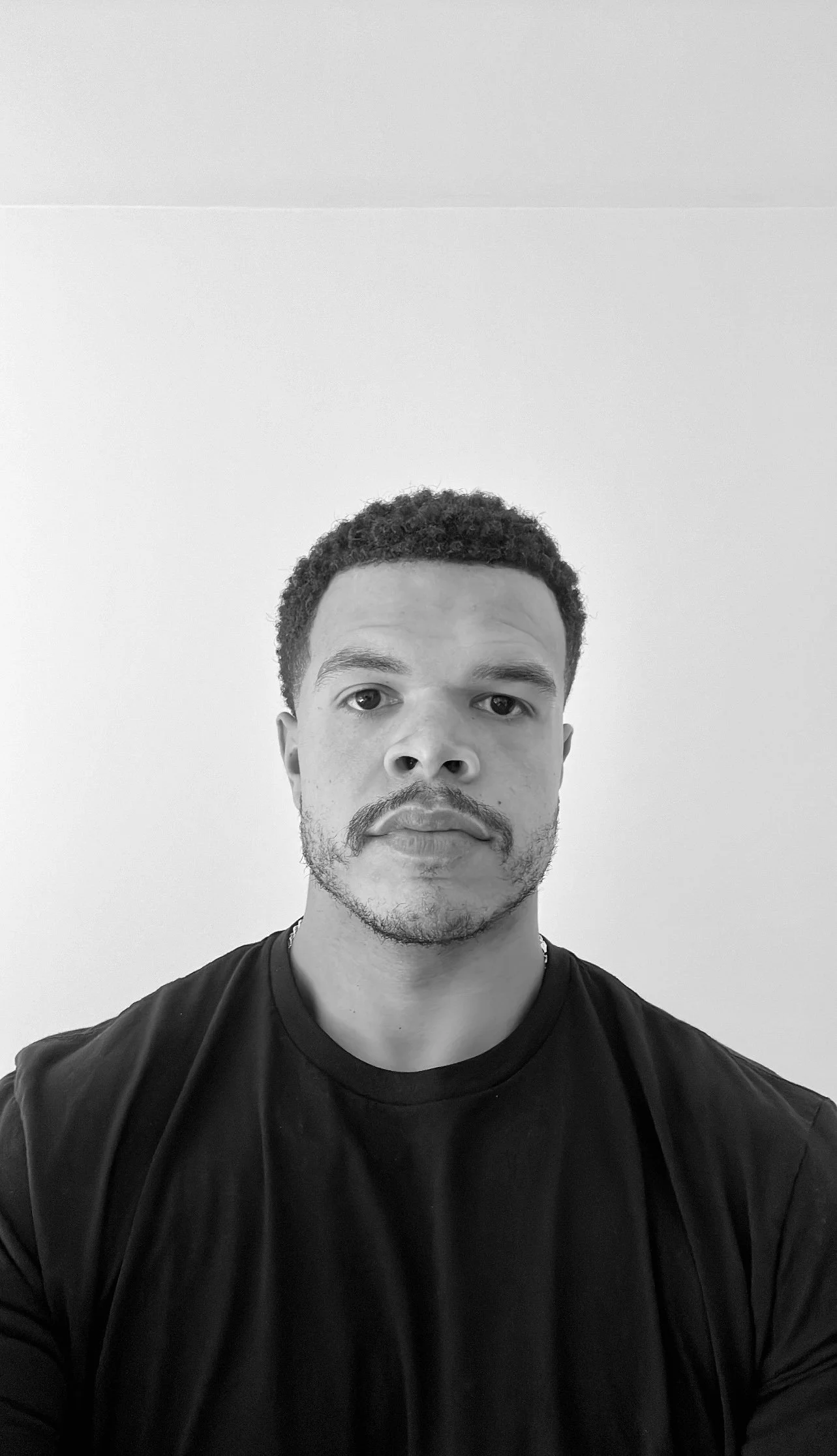 A black-and-white portrait of a young man with short curly hair, facial hair, wearing a black T-shirt, looking directly at the camera against a plain light-colored background.