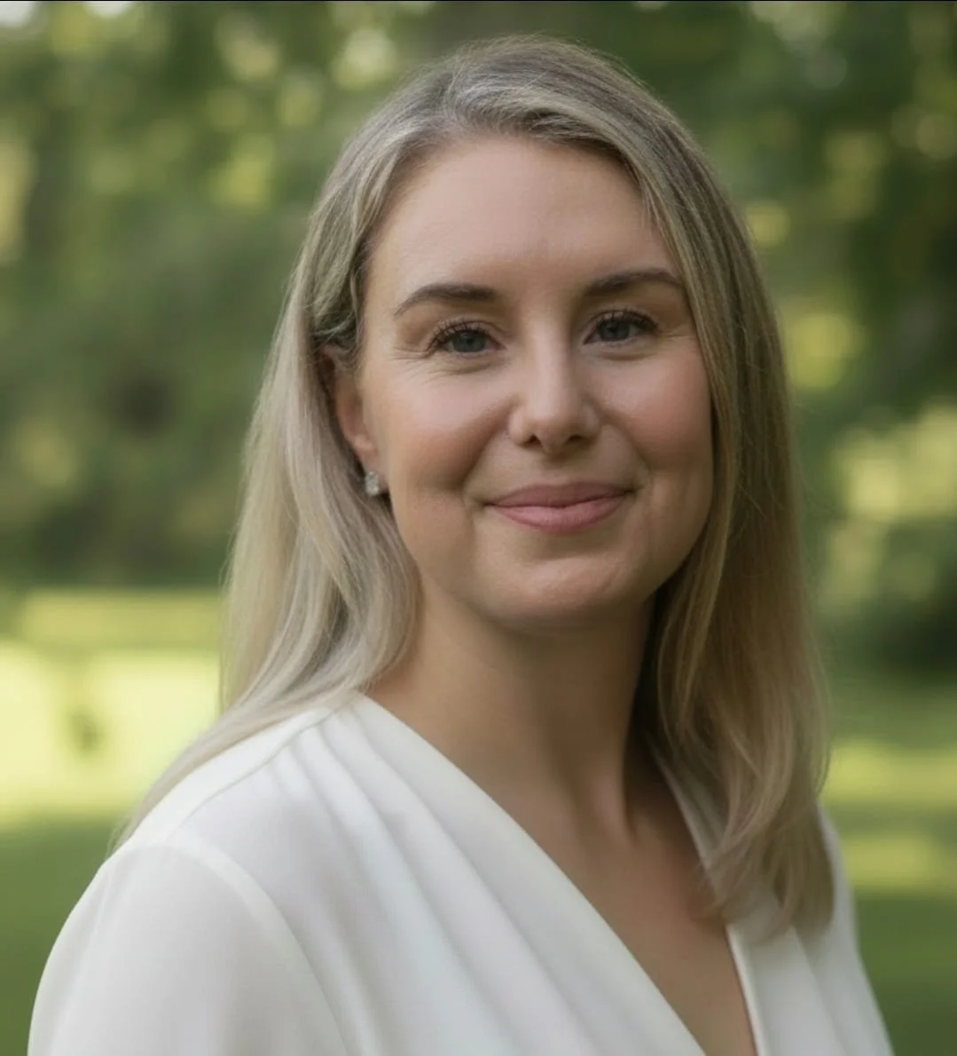 A woman with blonde hair, wearing a white blouse, standing outdoors with a blurred green background.