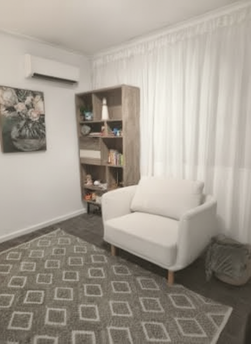 Living room interior with a white armchair, a wooden bookshelf filled with books and decorative items, a patterned rug, and a wall-mounted air conditioner, with white curtains covering the windows.