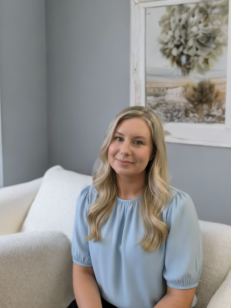 A young woman with long blonde hair and a light blue blouse sitting on a beige couch in a living room. She is smiling softly at the camera with a seaside landscape photo hanging on the wall behind her.