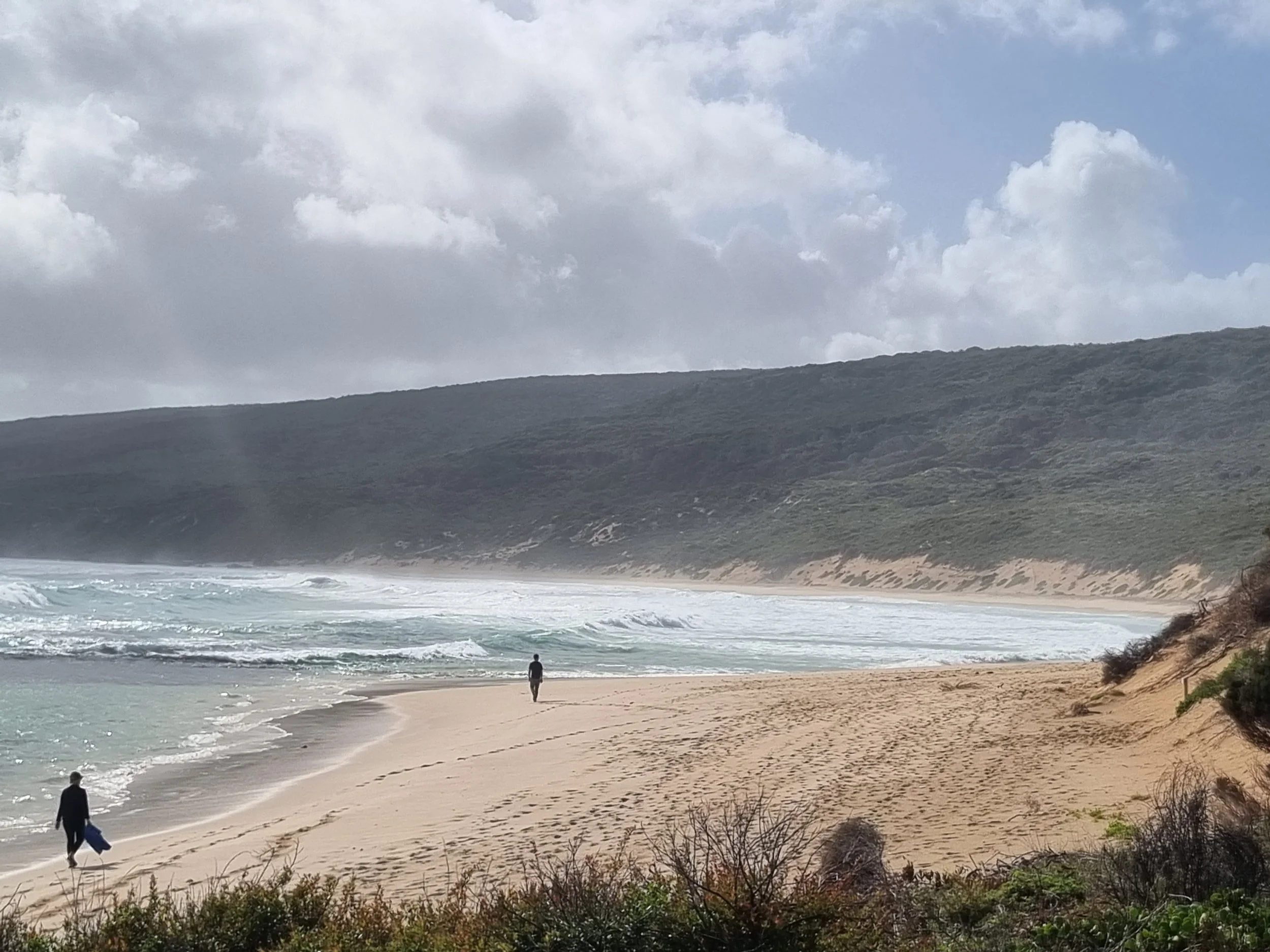 A sandy beach with two people, one carrying a surfboard, walking along the shoreline. The ocean has small waves, and there are hills in the background under a cloudy sky.