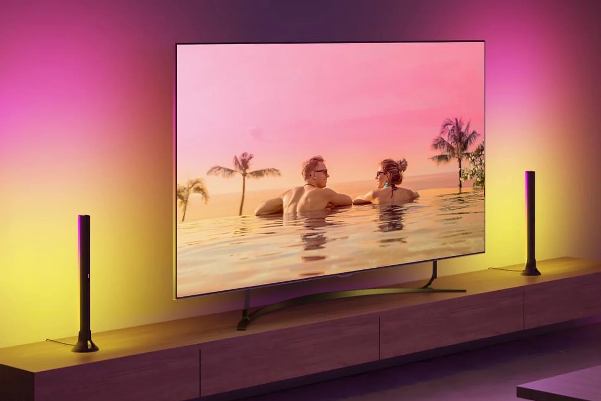 A modern flat-screen television displaying a romantic scene of a man and woman in a swimming pool with pink sunset sky and palm trees in the background, placed on a wooden stand with colorful ambient lighting.