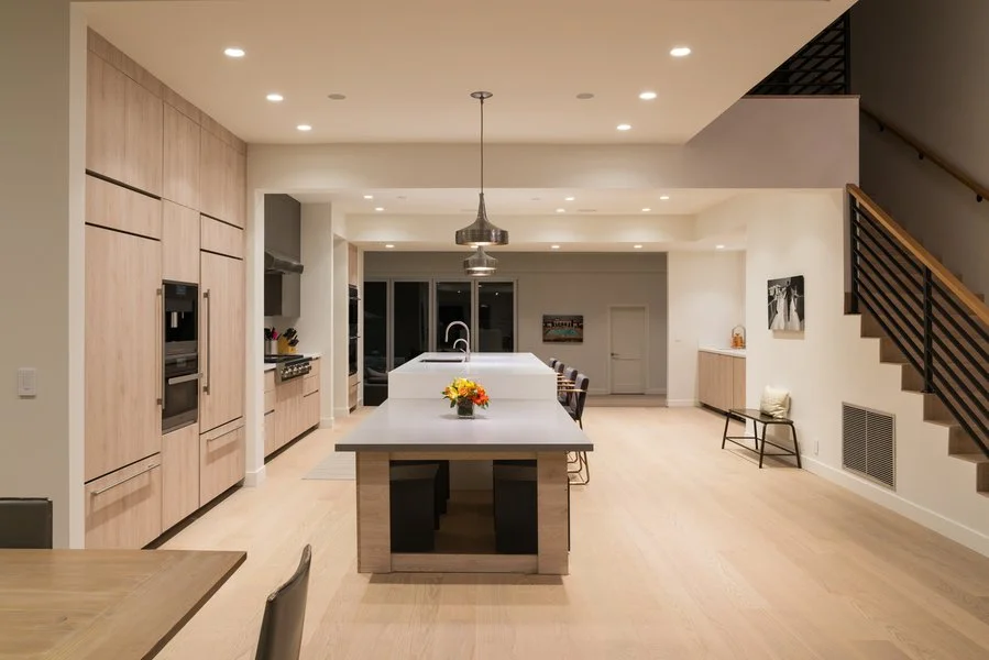 Modern open-concept kitchen with light wood cabinetry, a large center island with bar stools, built-in appliances, and a staircase with black railing leading upstairs.