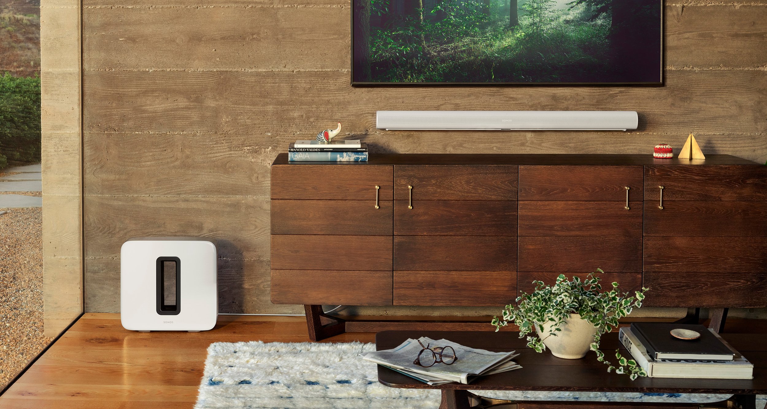 Living room sideboard with a potted plant, books, decorative items, and a wall-mounted television above, with an abstract painting on a wooden wall and a white subwoofer on the brown hardwood floor.