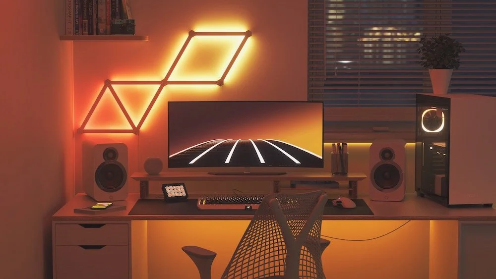Modern gaming or work desk setup with a computer monitor displaying a retro stylized landscape, surrounded by speakers, a keyboard, mouse, and decorative lighting, including a neon geometric wall art and warm ambient lights.
